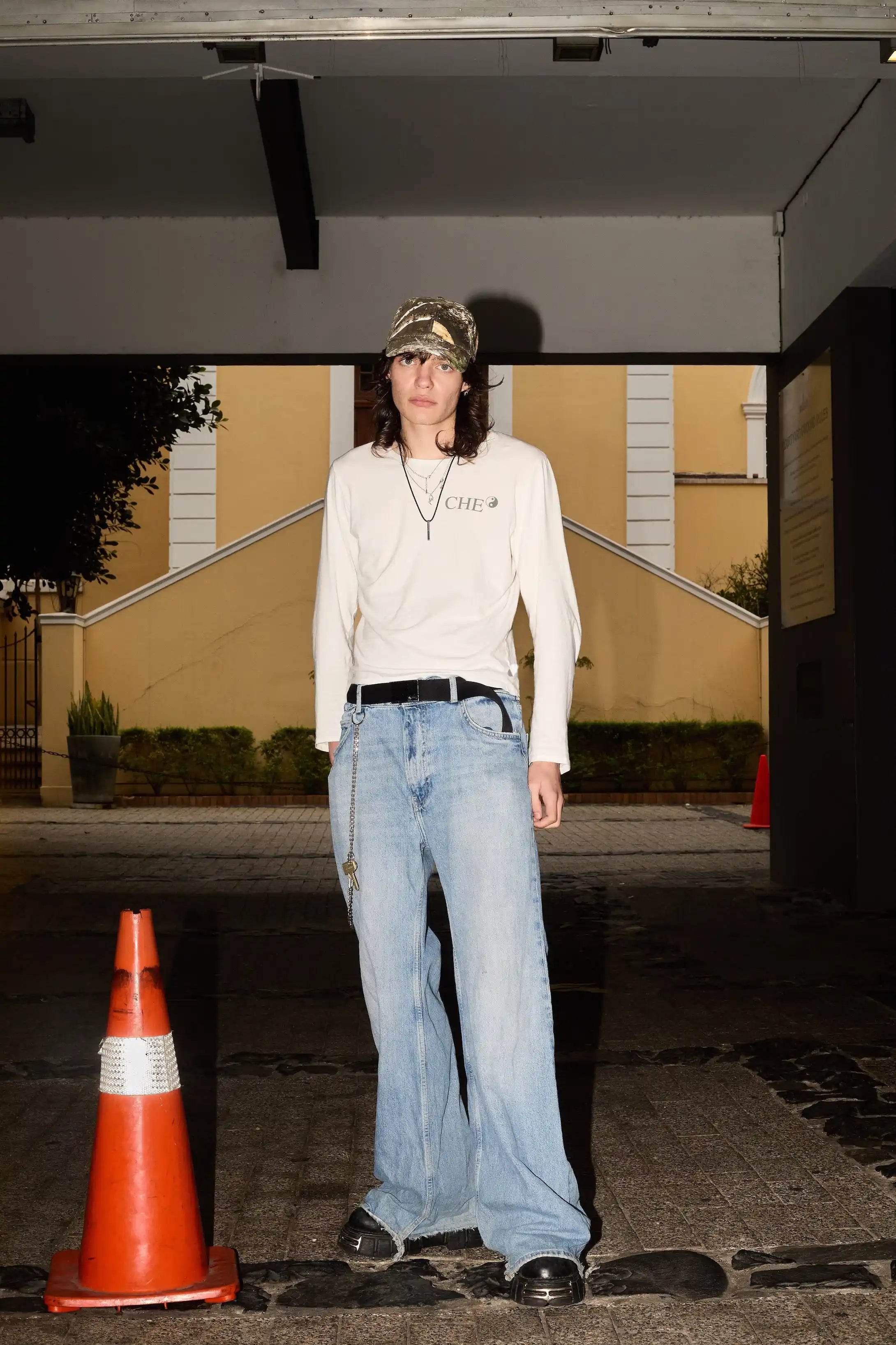 Person wearing a beige sweater and blue jeans standing next to traffic cones on a street.