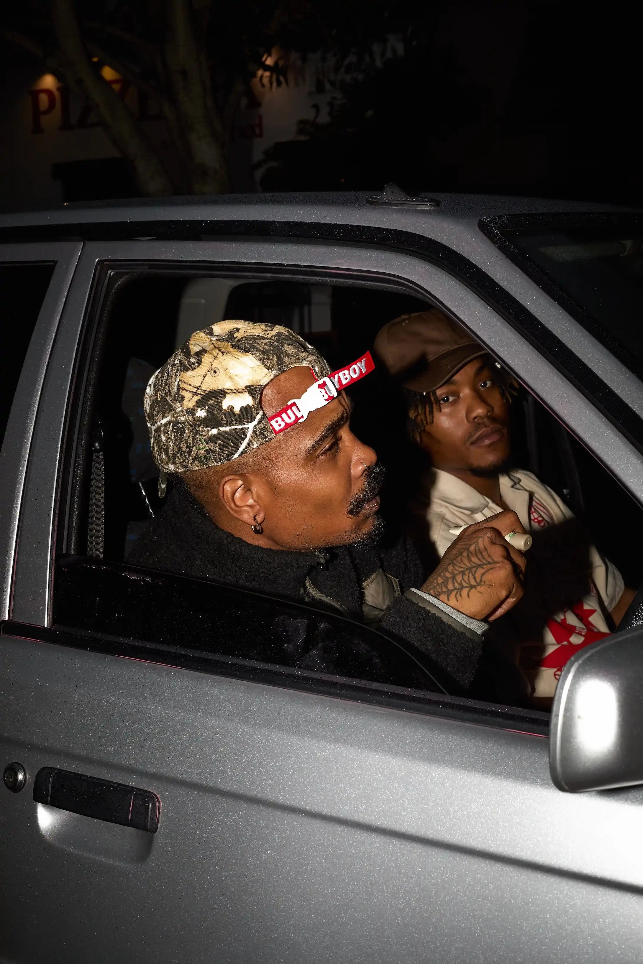 Two men sitting in a car at night, one wearing a camouflage cap with a visible brand logo.