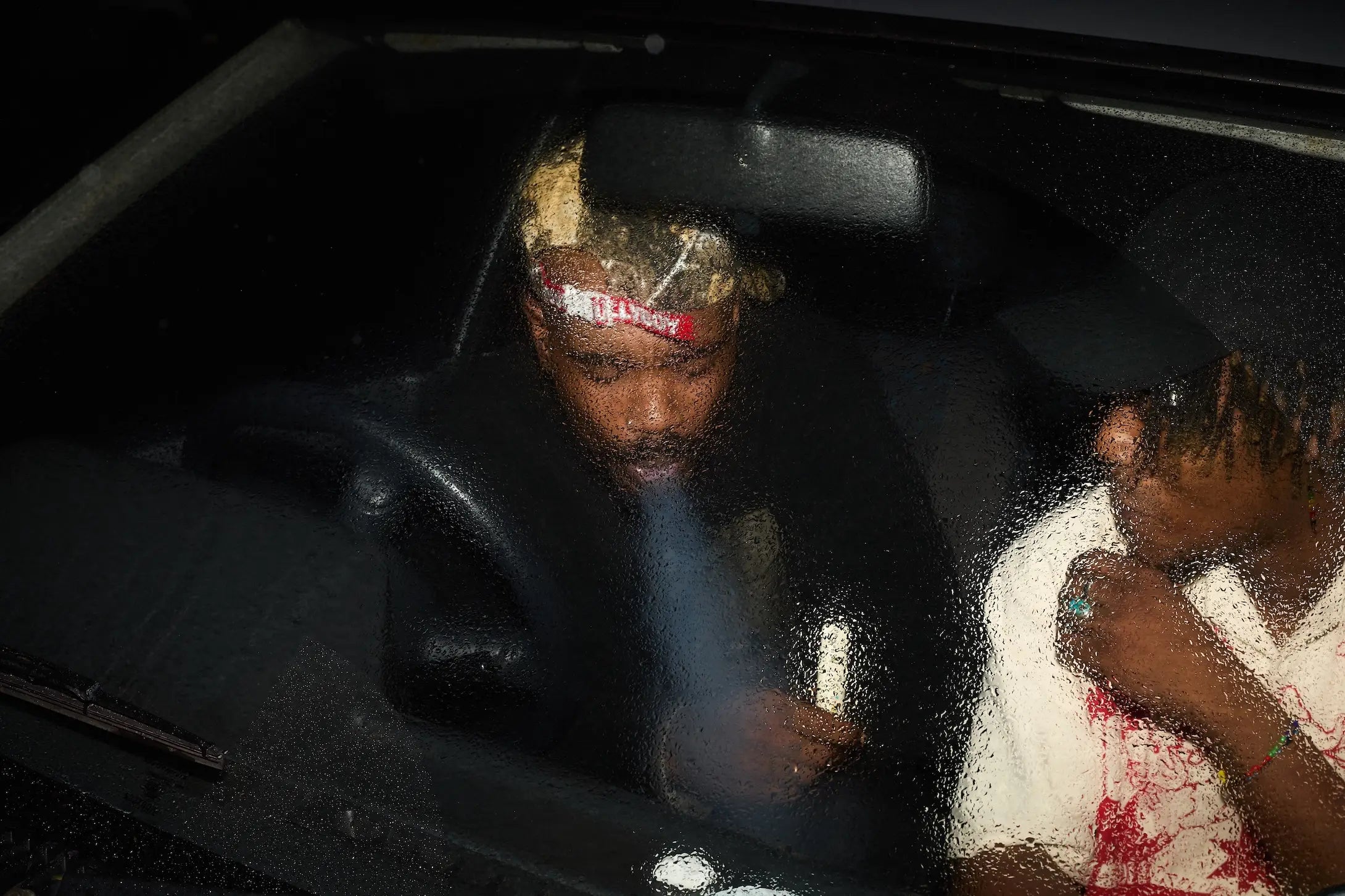 Person sitting inside a vehicle with a child, both smoking, in a dark setting.