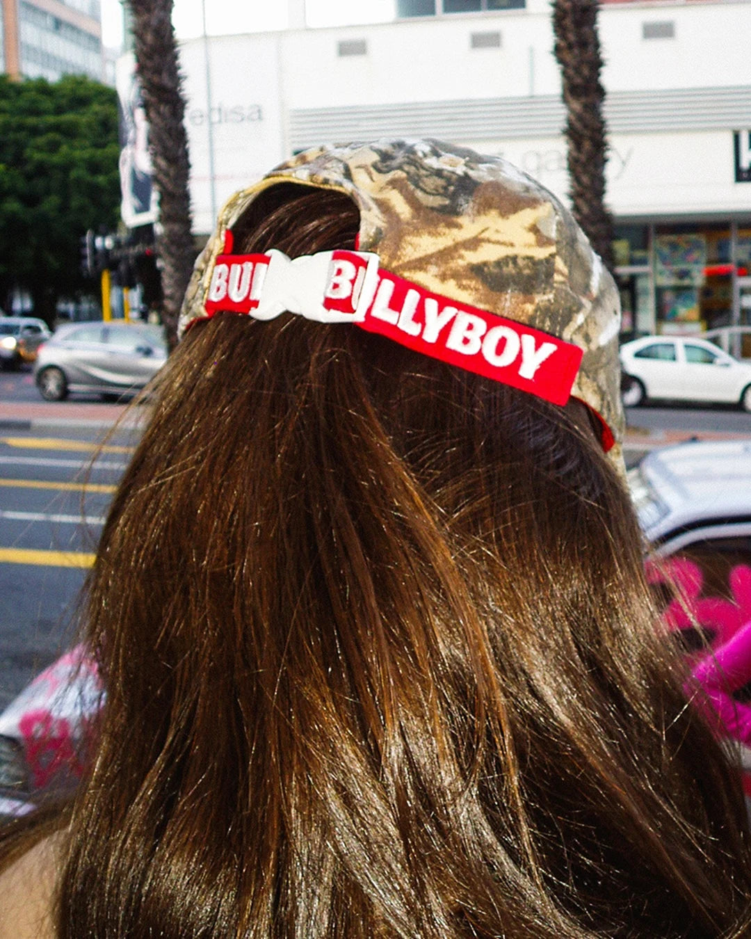 Person wearing a camouflage cap with a red 'Bully Boy' headband on a city street.