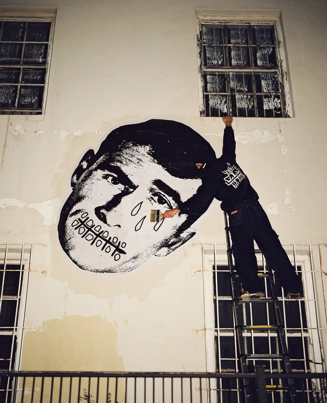 Person on a ladder painting a large wheat paste mural of a face on a wall.