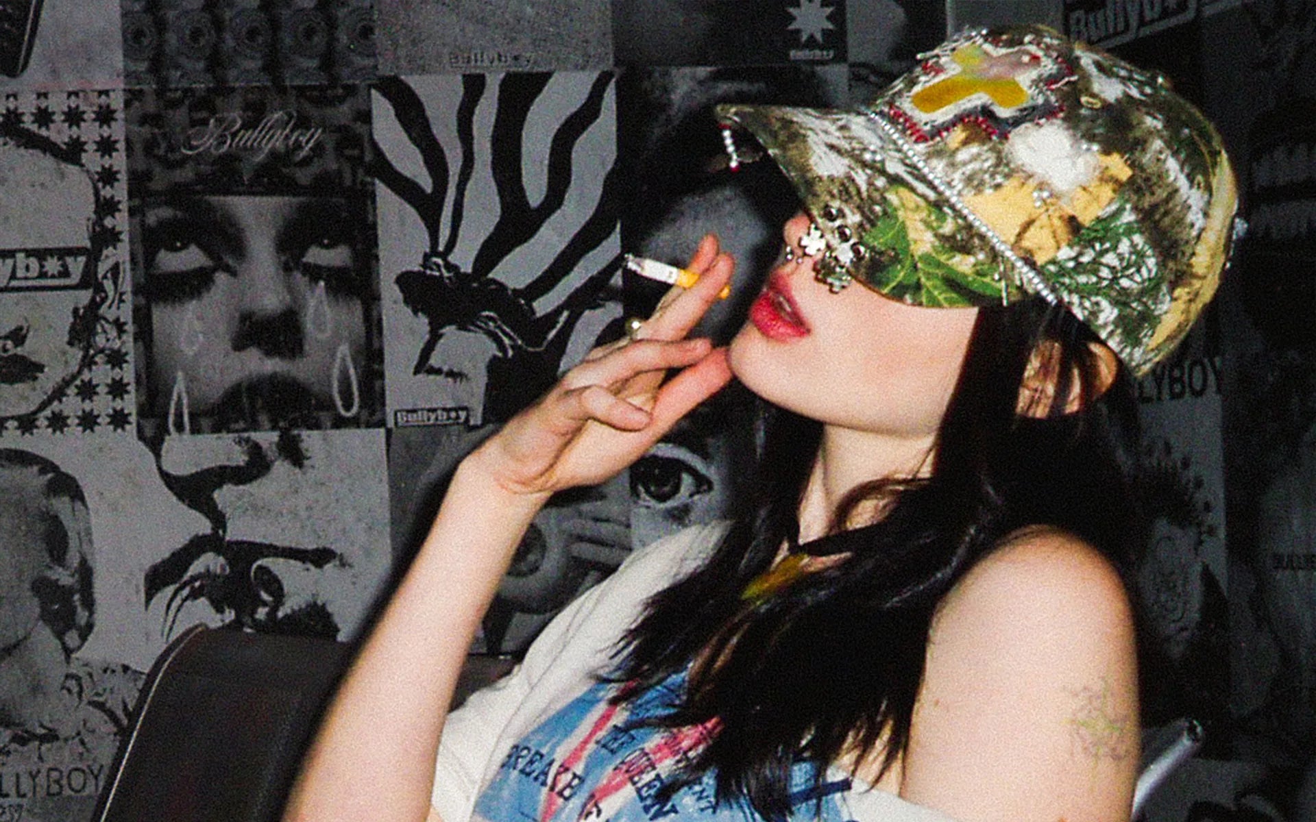 Person applying makeup with a camouflage-patterned hat in front of a graphic wall.