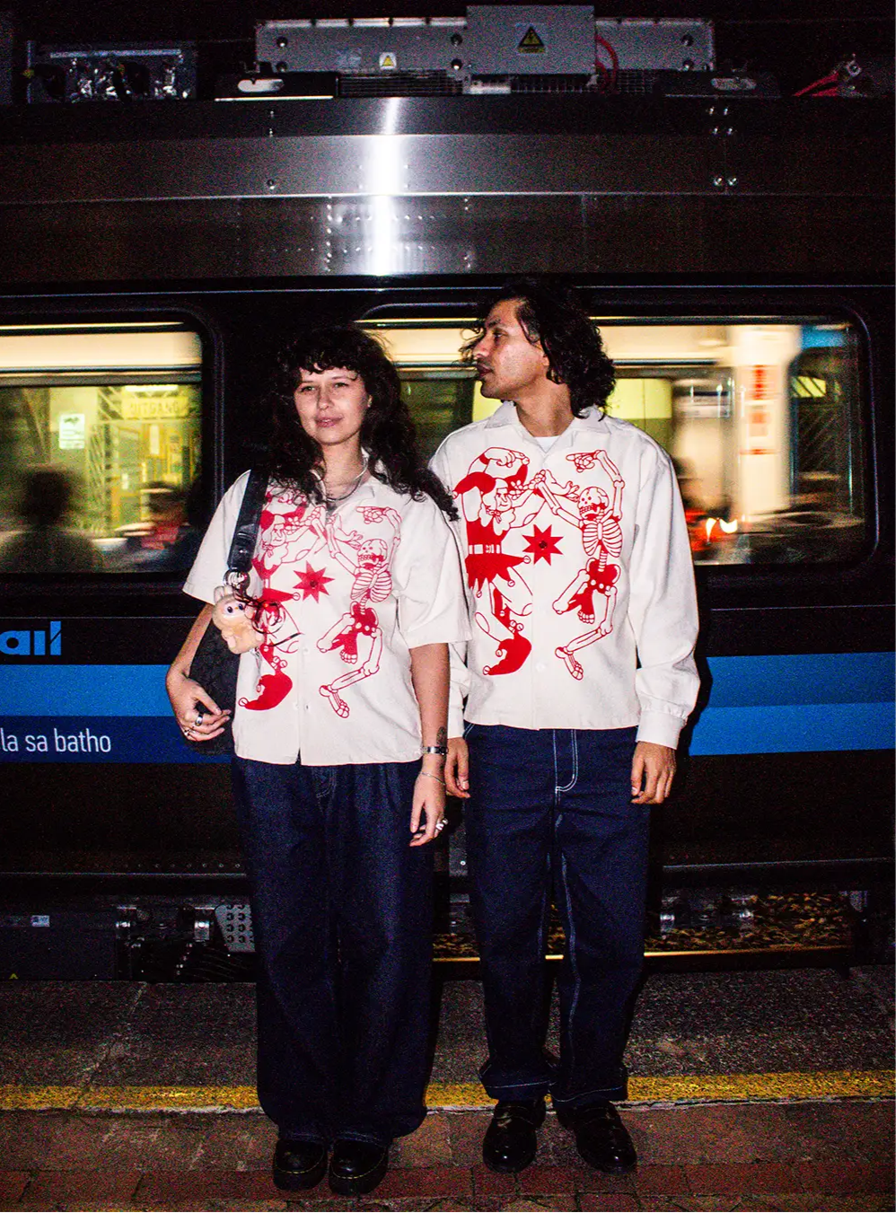A trendy couple stands near the Cape Town train tracks at night while wearing the new BullyWopp button up shirts