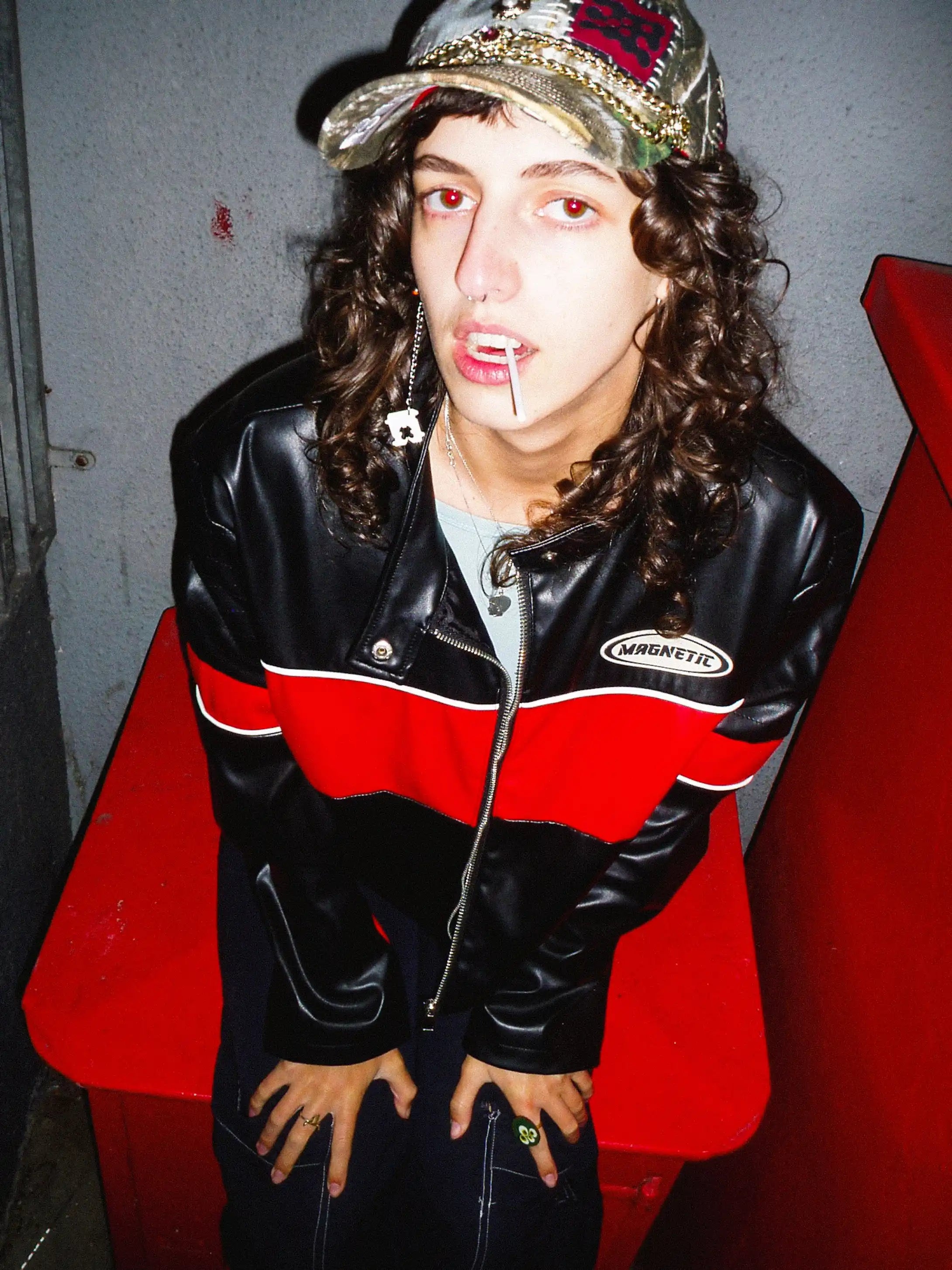 Person wearing a black and red jacket with a cap, sitting against a red and gray wall.