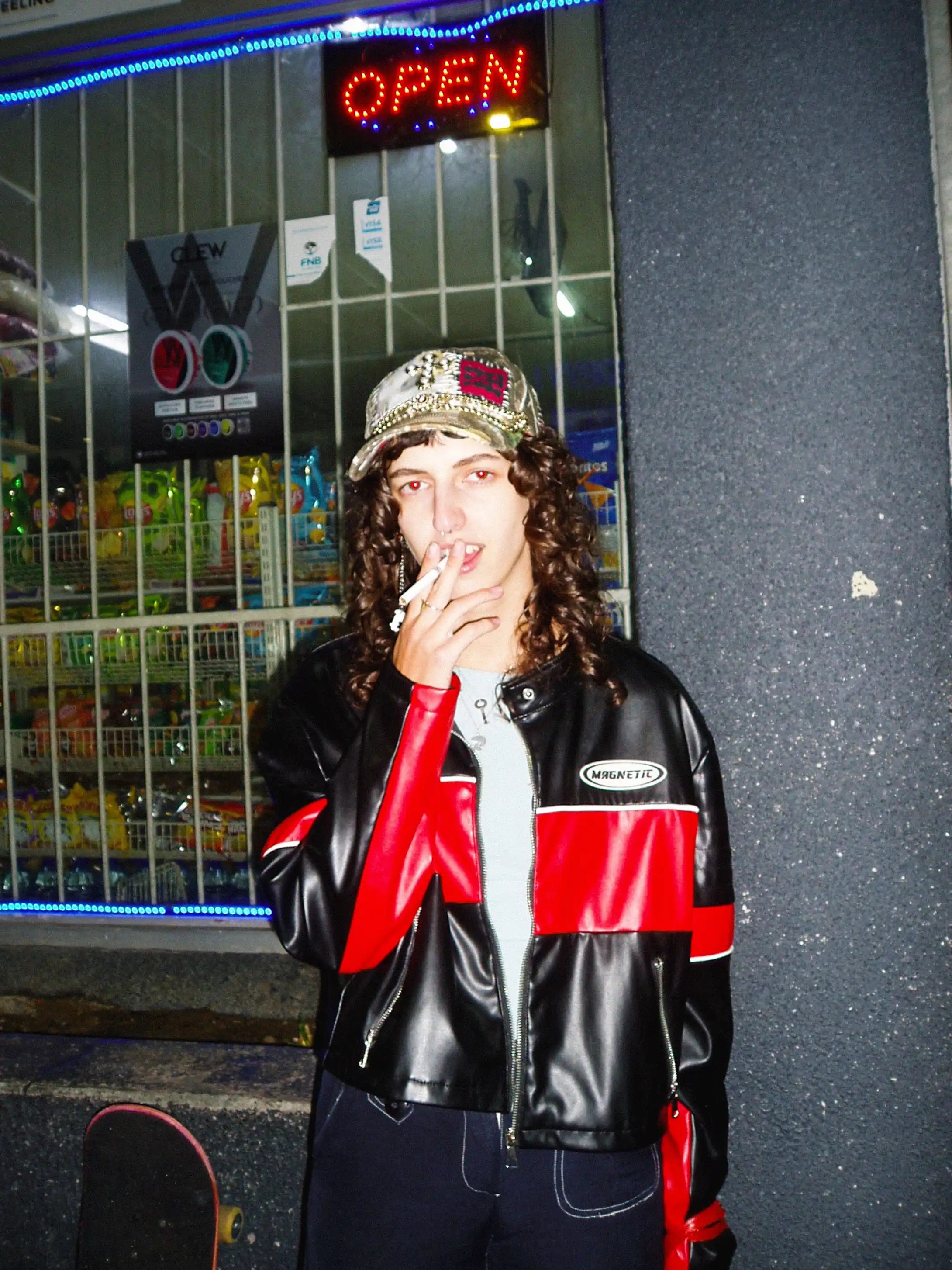 Person wearing a black and red jacket with a cap, standing in front of a store with an 'Open' sign.