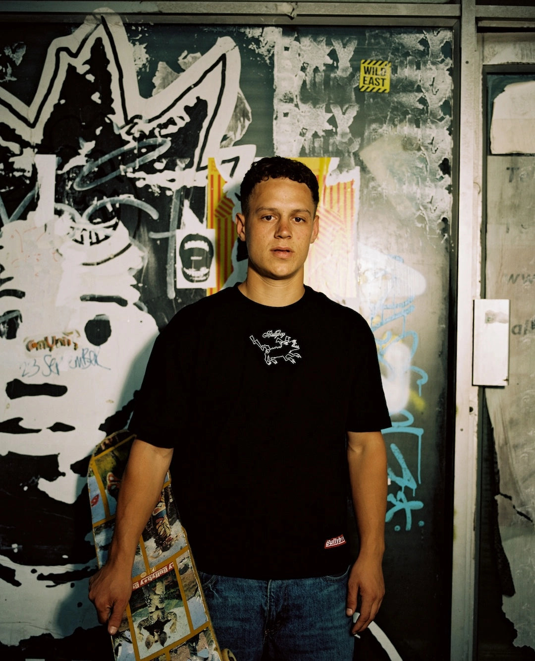Person wearing a black t-shirt with a logo in front of a graffiti-covered wall.
