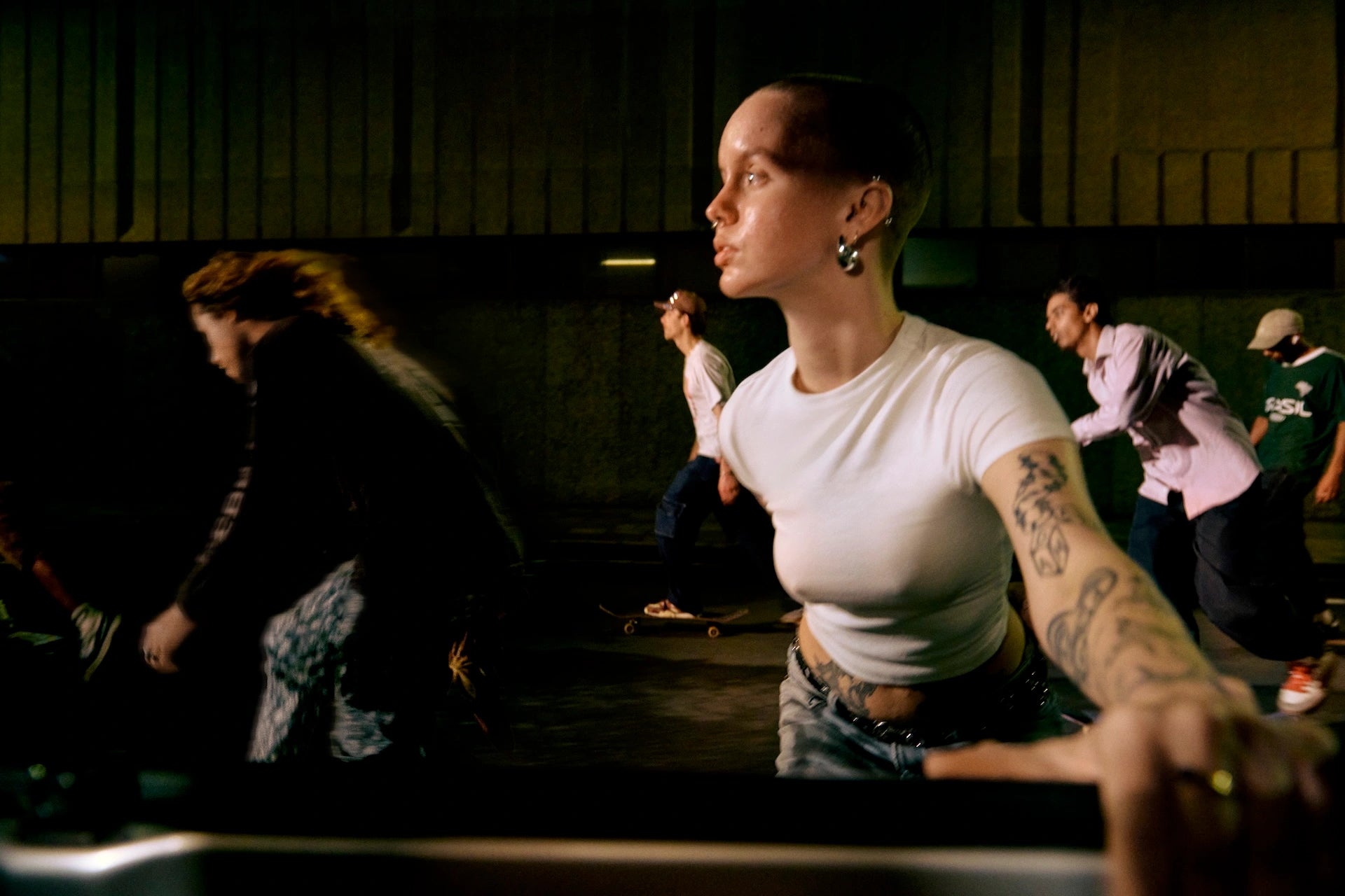 Person with tattoos sitting in a dimly lit room with other people in the background