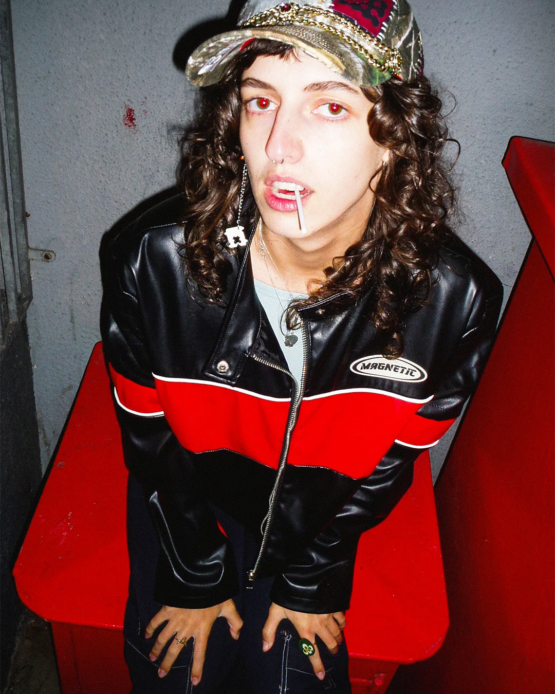 Person wearing a black and red jacket with a gold and red cap, sitting on a red bench against a gray wall.