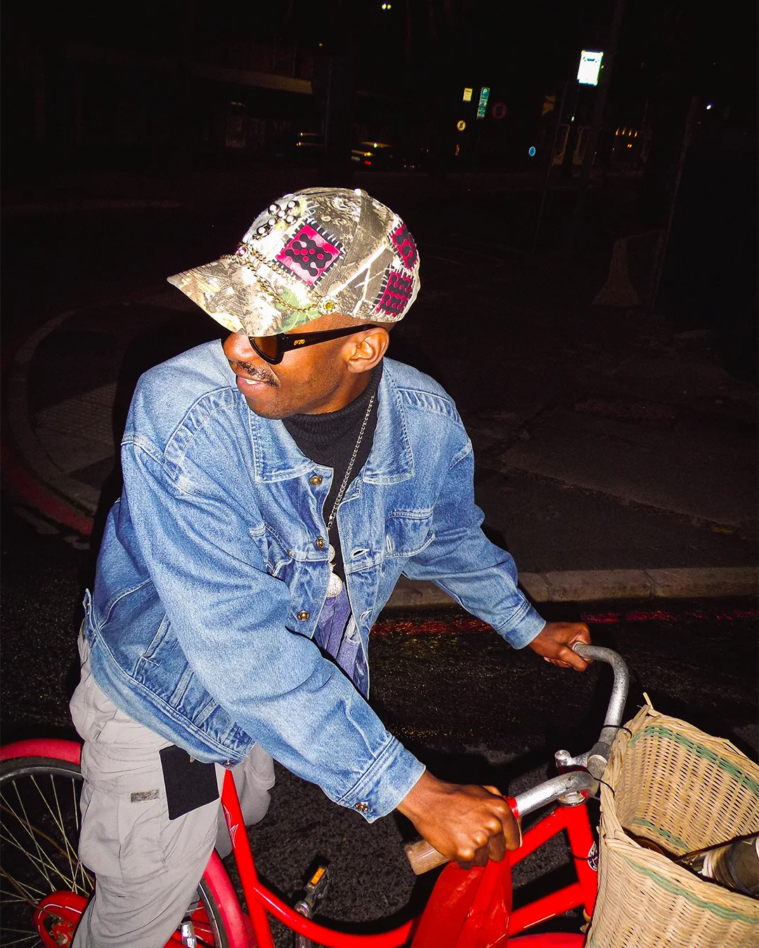 Man wearing a denim jacket and colorful cap riding a red bicycle at night.