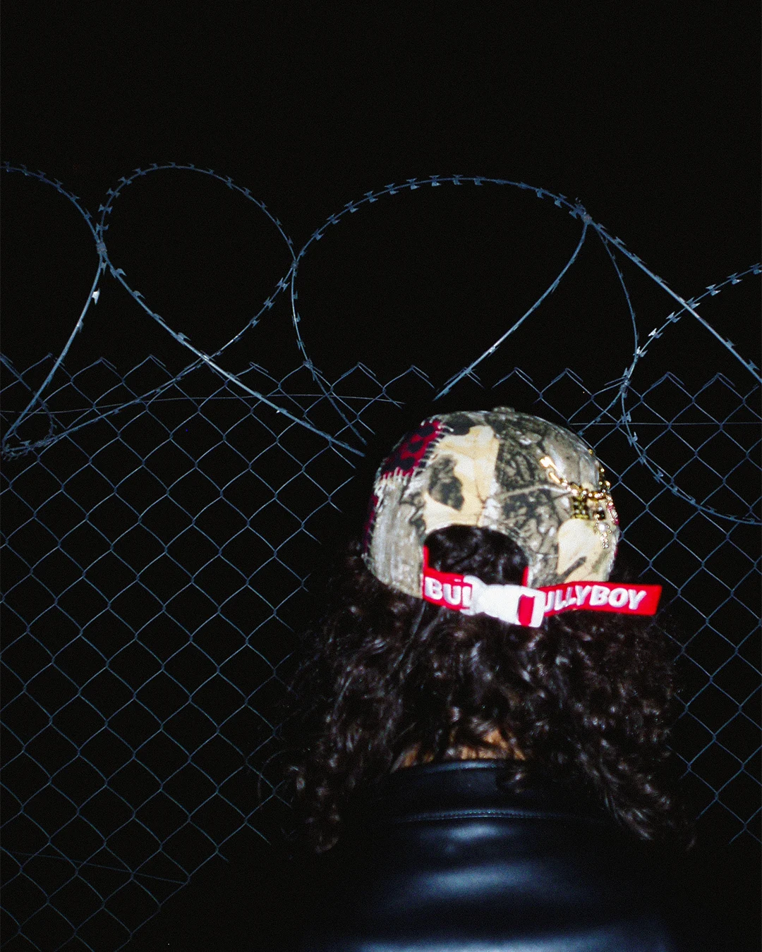 Person wearing a cap with 'Bullyboy' logo against a barbed wire fence background