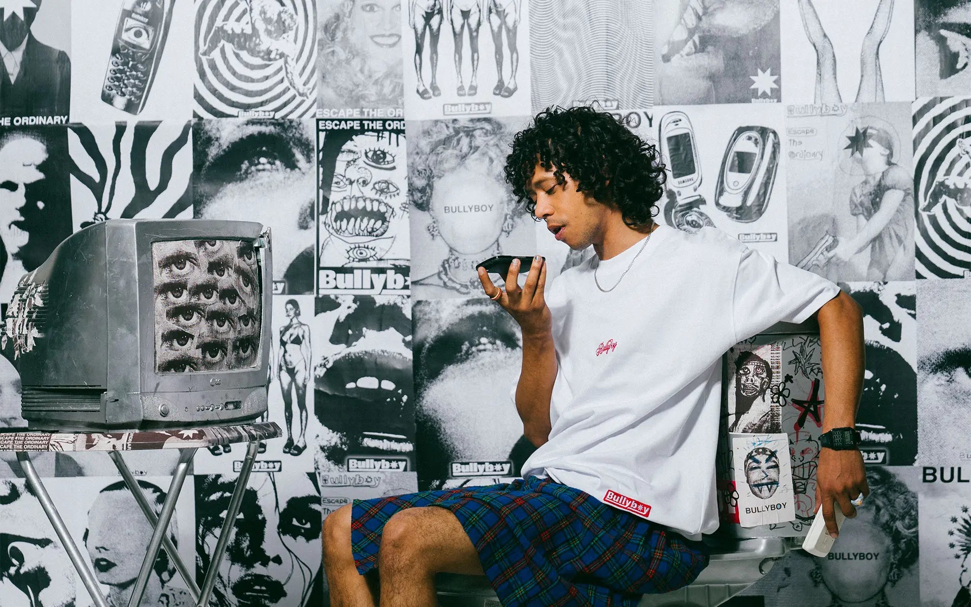 Person sitting against a wall with graphic posters, wearing a white t-shirt and plaid shorts.