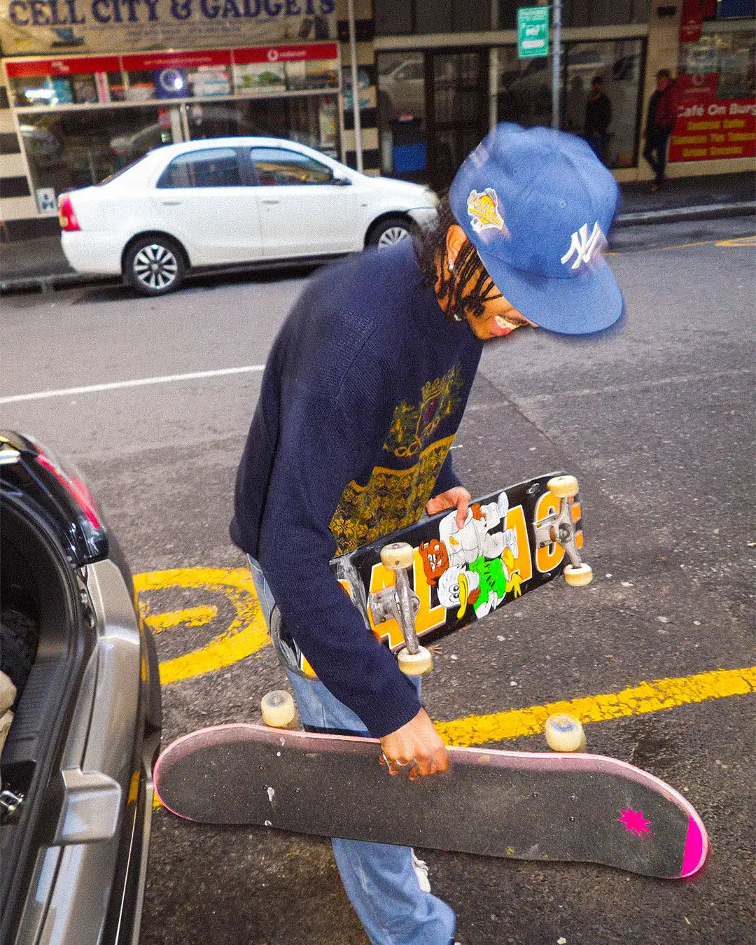 Person holding a skateboard on a city street with a car and store in the background