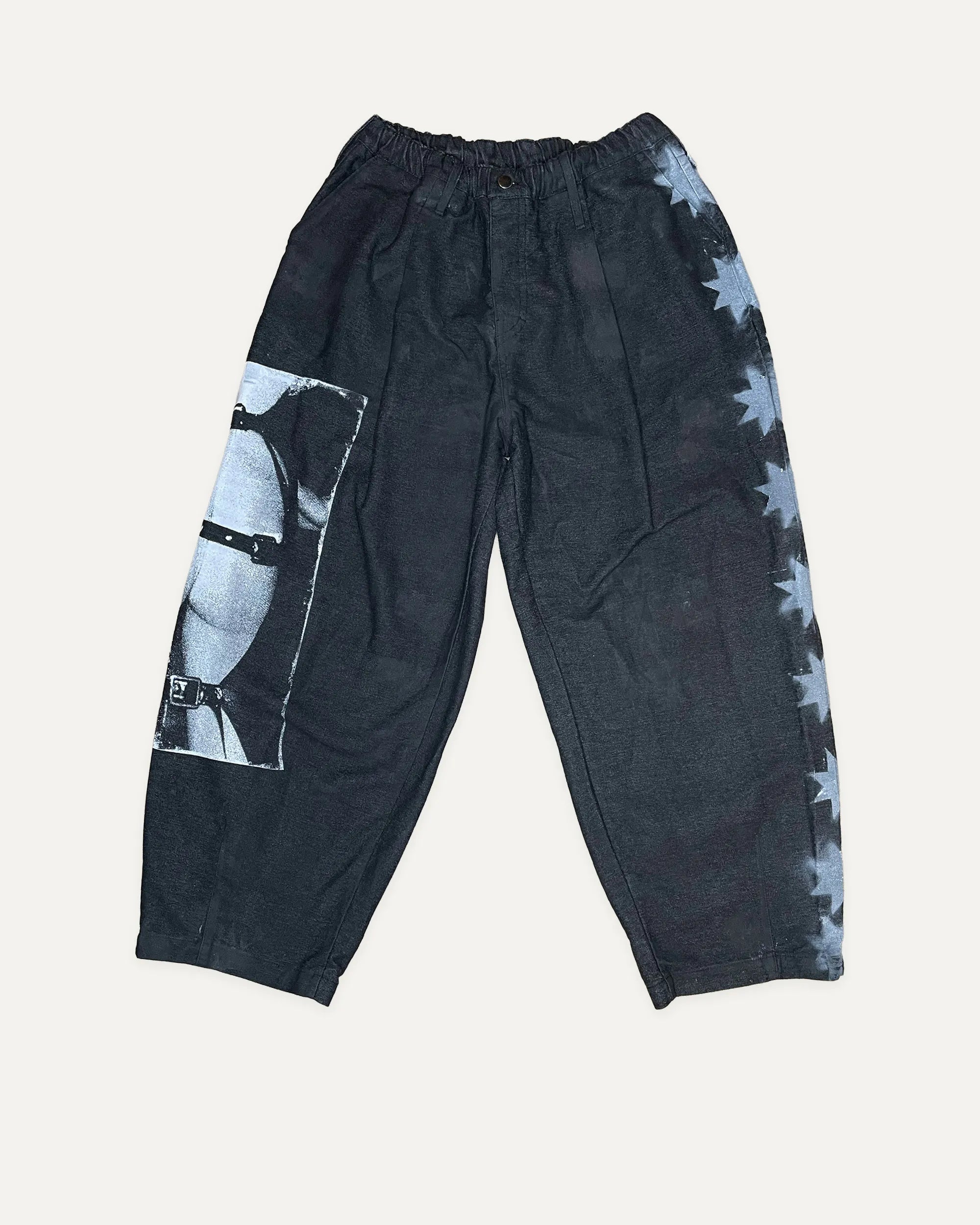 Black washed cotton pants featuring a large black-and-white graphic print on the right leg and a side pattern of light blue stars, laid flat on a light beige background.