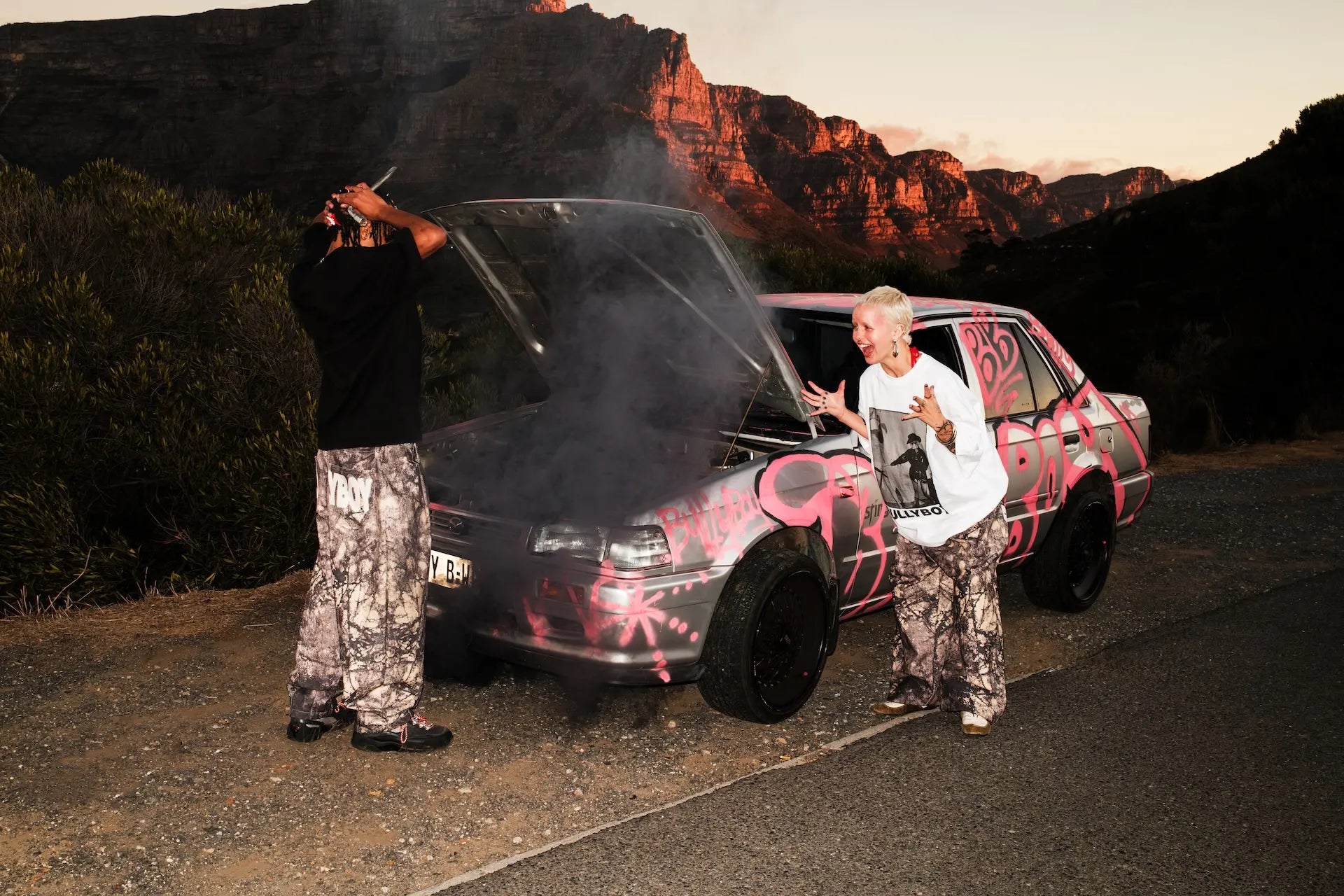 Two people standing next to a car with pink and black design in a mountainous area.
