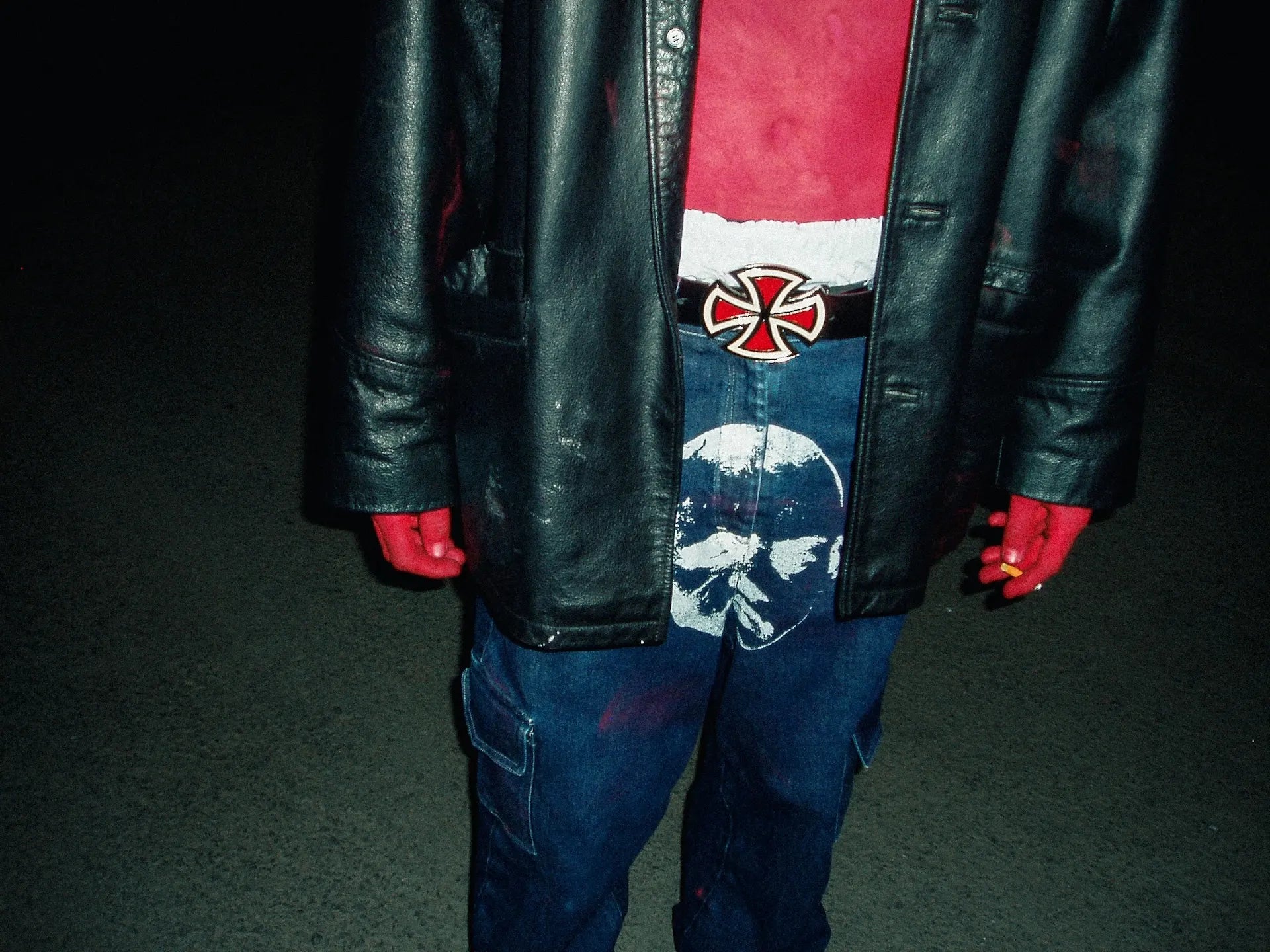 Person wearing a black leather jacket, blue jeans with a graphic design, and red gloves on a dark background.