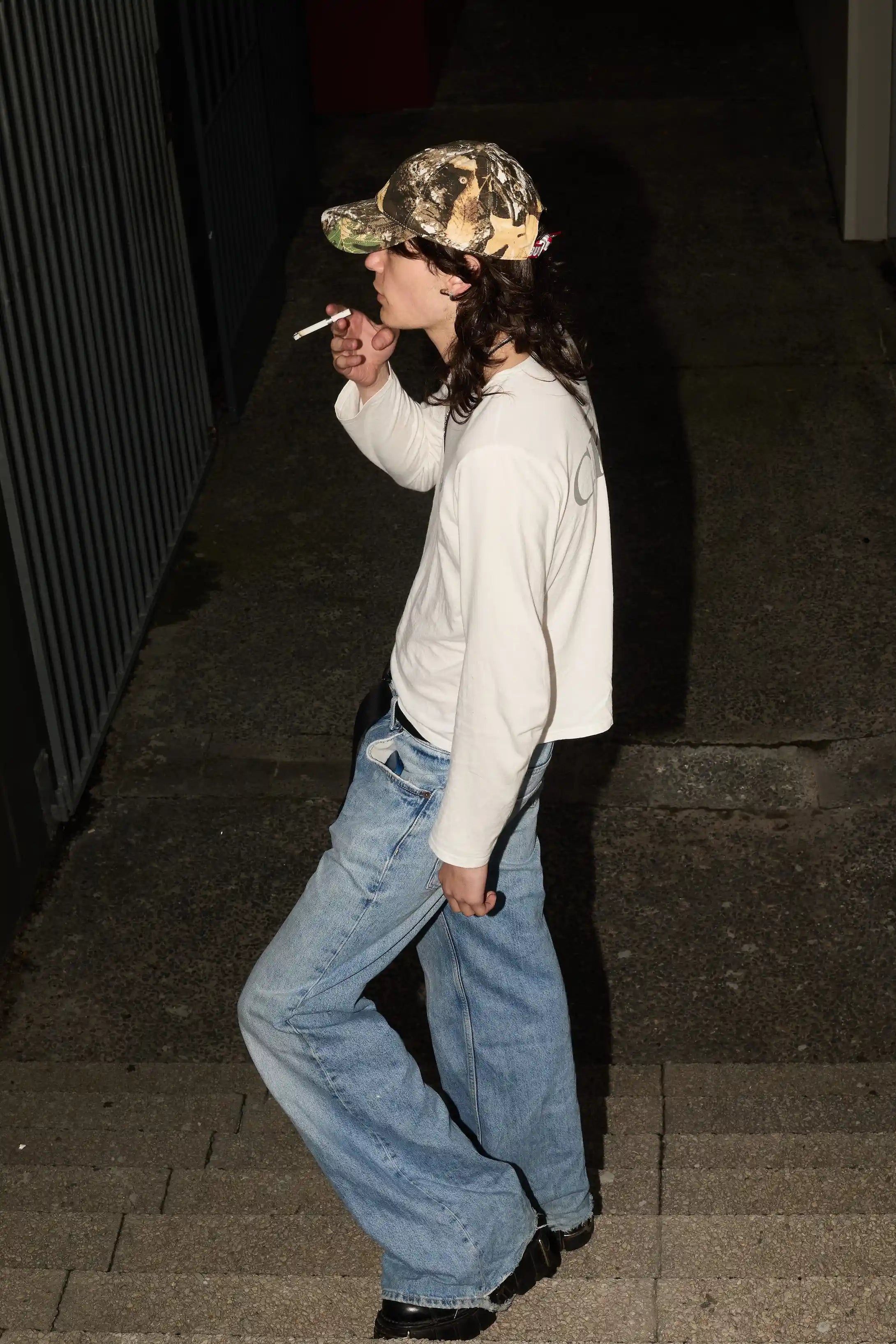 Person wearing a camouflage cap and light-colored sweatshirt, smoking on a street.