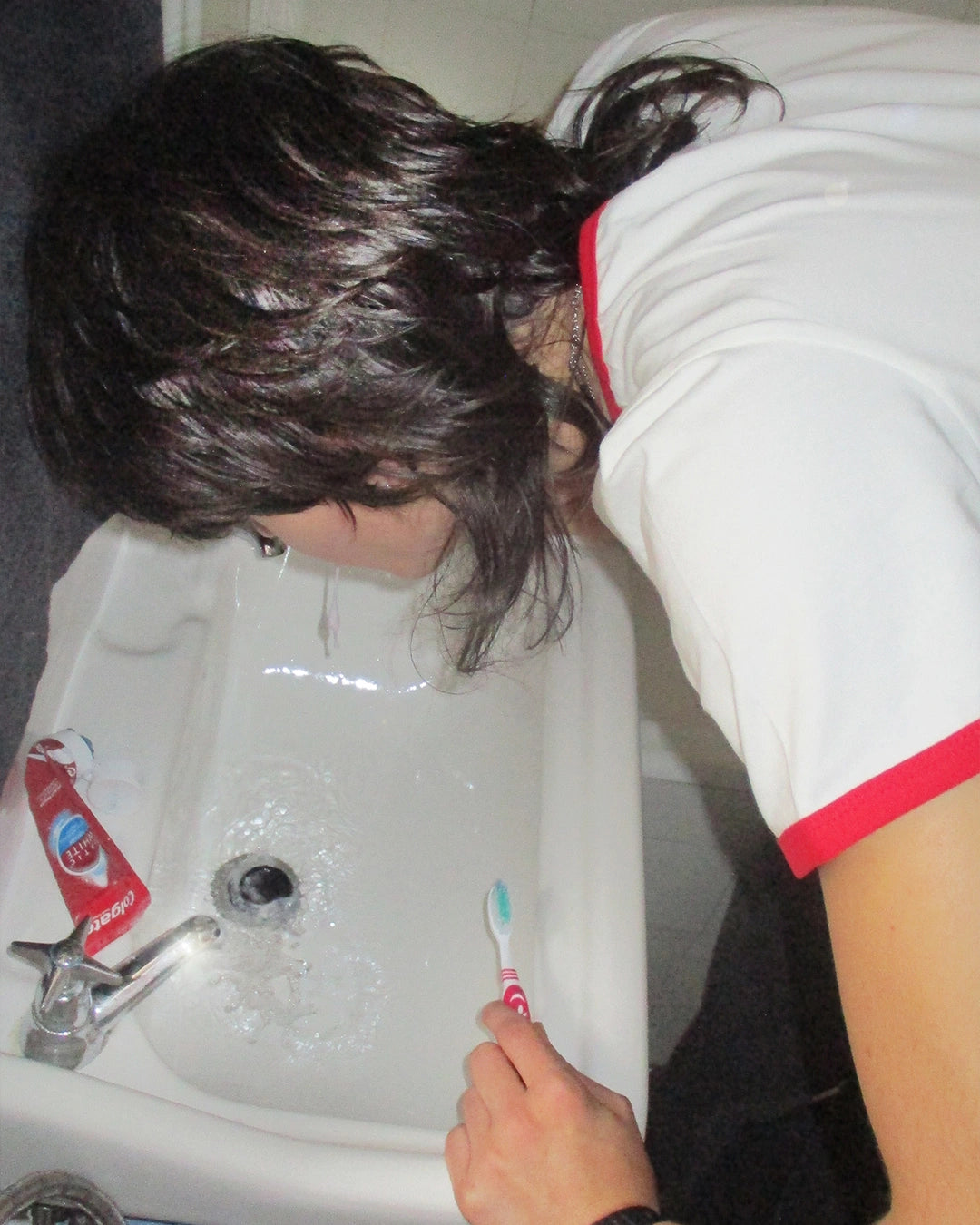 Person leaning over a sink with a toothbrush, wearing a white shirt with red trim.