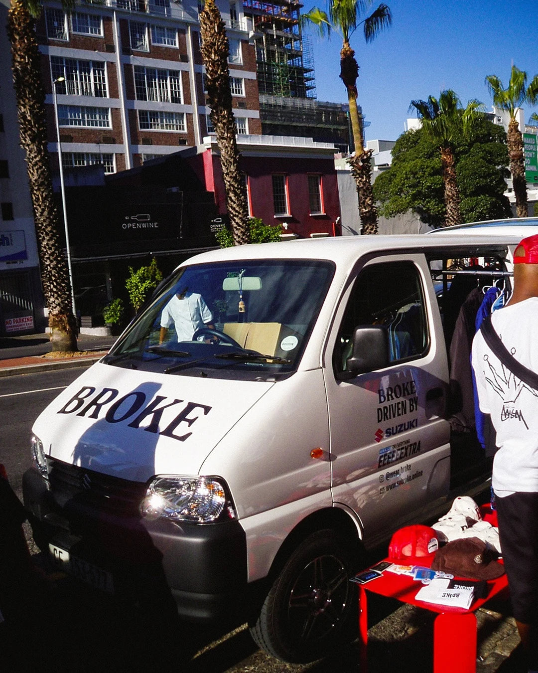Small white van with 'BROKE' branding on a city street with palm trees and buildings.