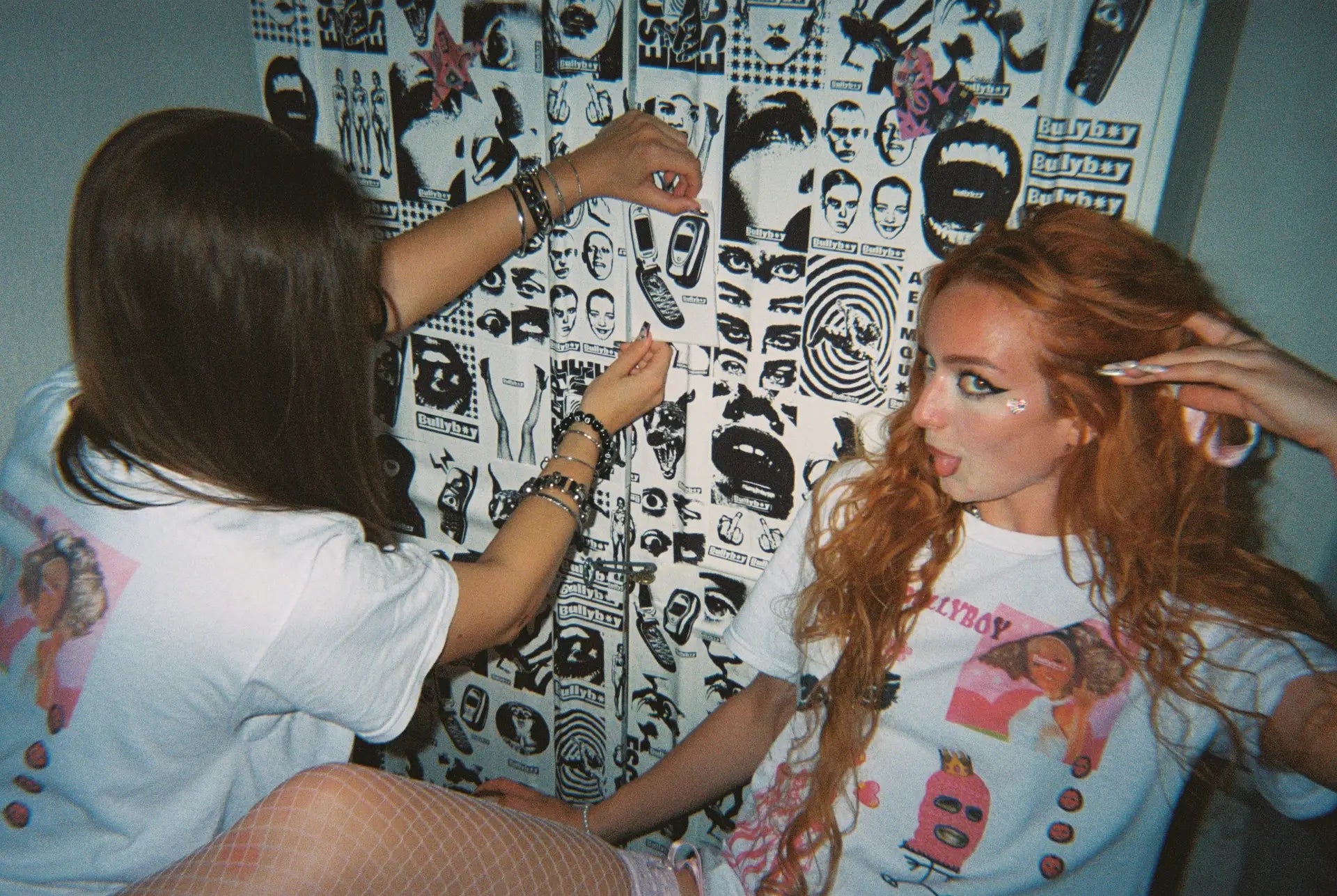 Two women sitting together with a wall covered in stickers.