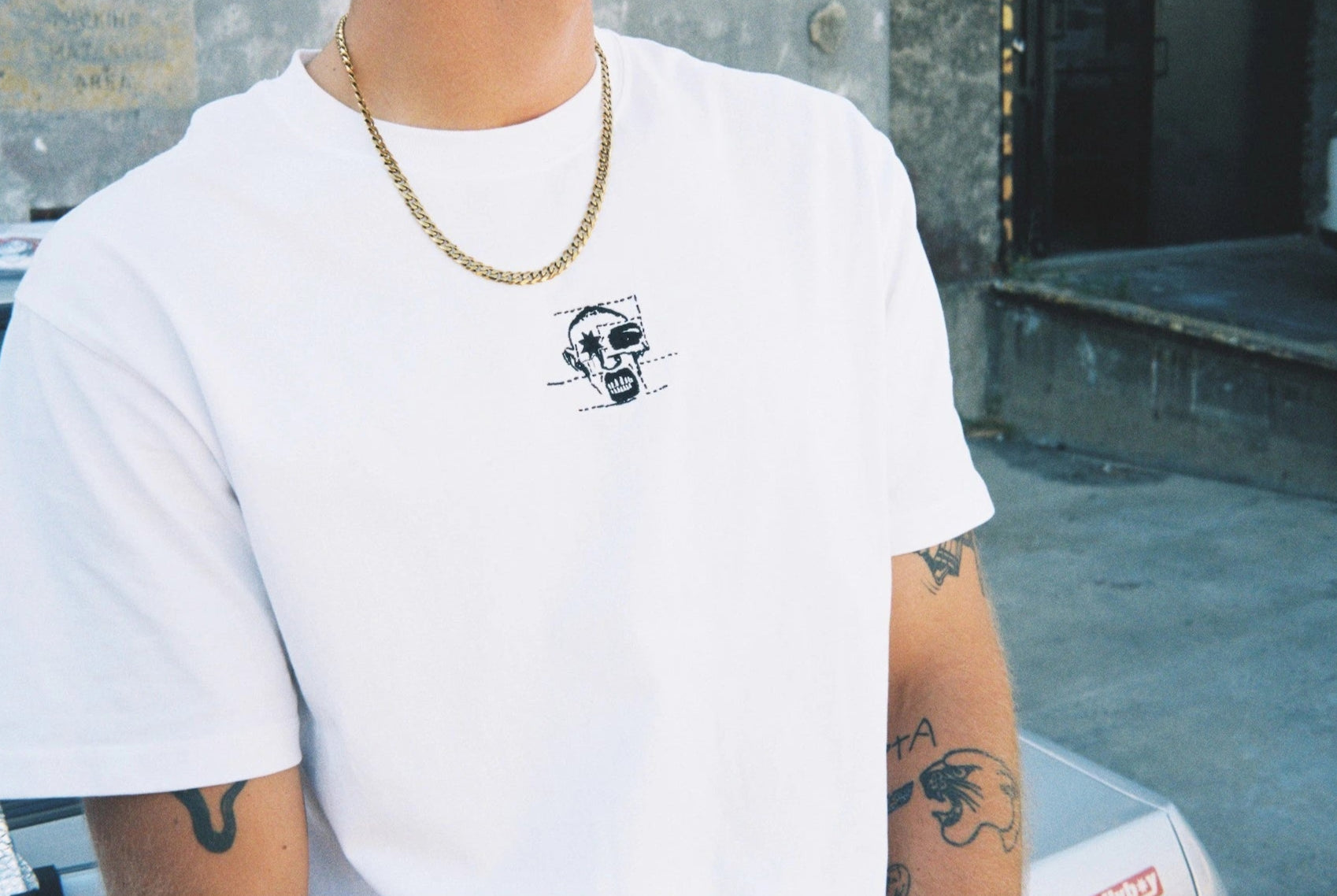 Person wearing a white t-shirt with a small graphic design, standing in an urban setting.