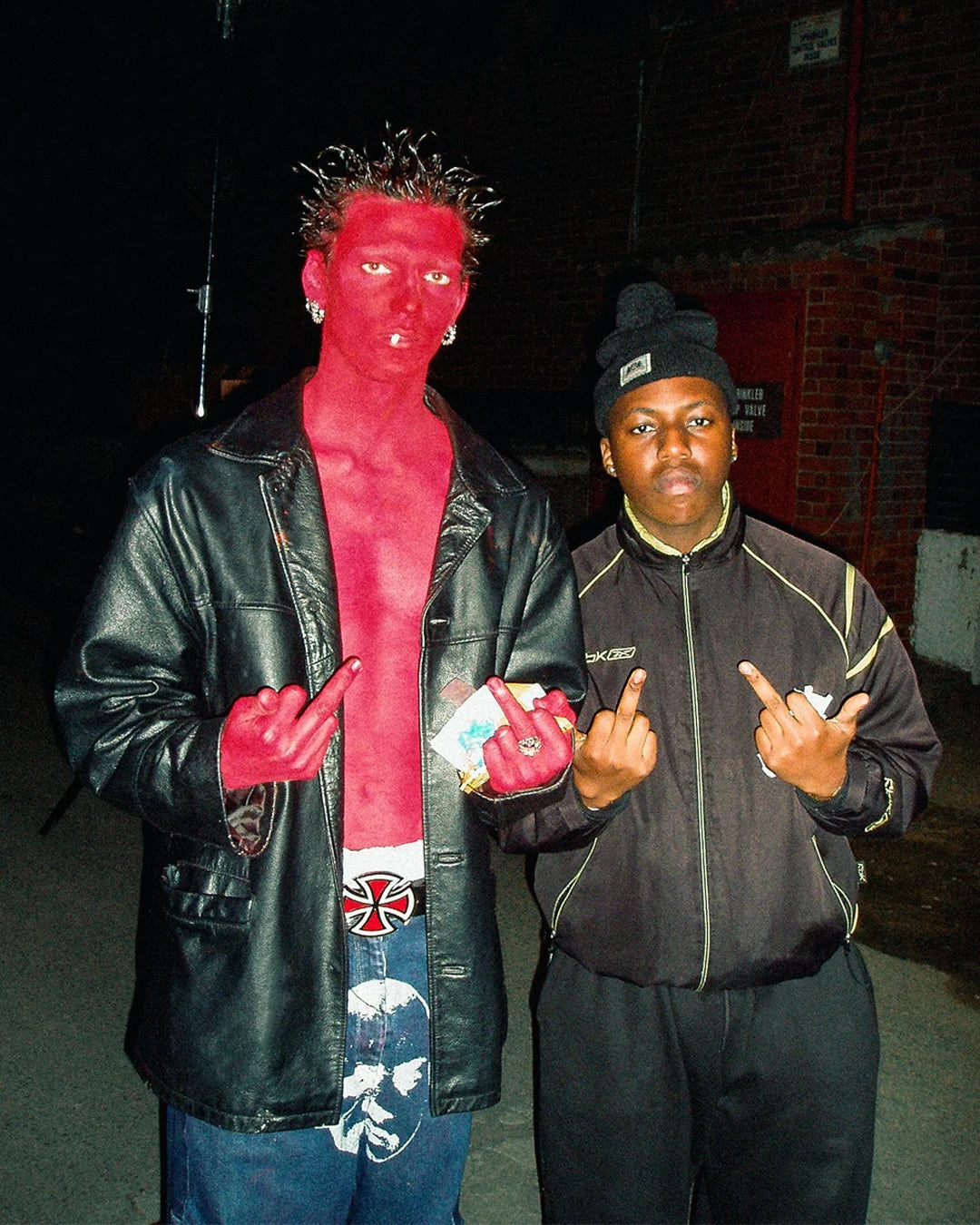 Two people posing together with one person wearing a red face paint and black leather jacket.