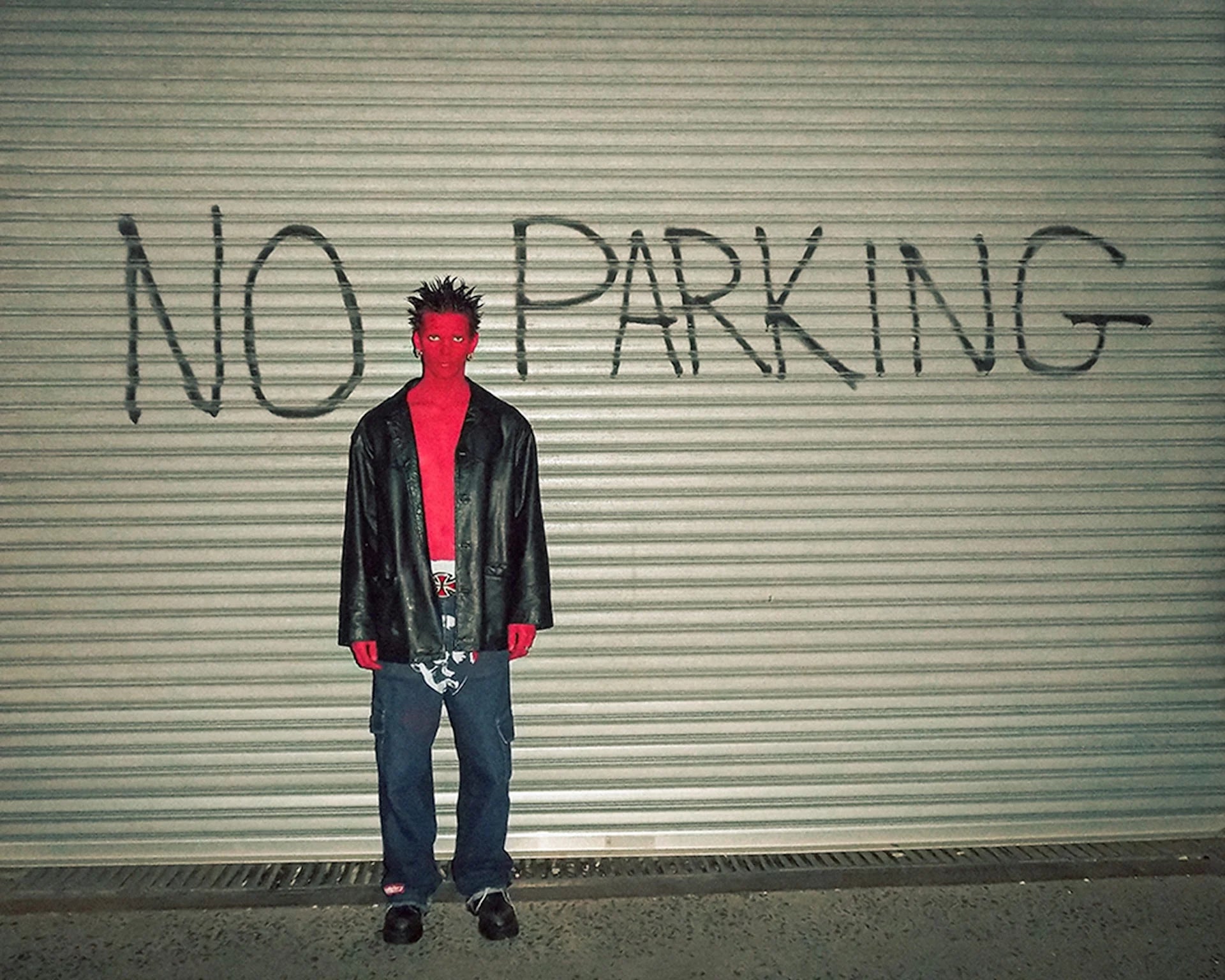 Person wearing a black leather jacket and red shirt standing in front of a 'No Parking' sign on a metal shutter.