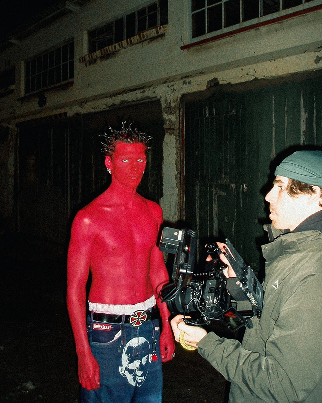 Person with red body paint being filmed by a cameraman in an industrial setting