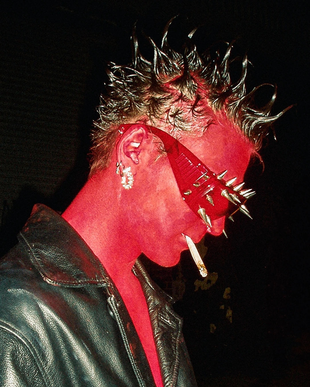 Person wearing a red mask with spikes and leather jacket against a dark background