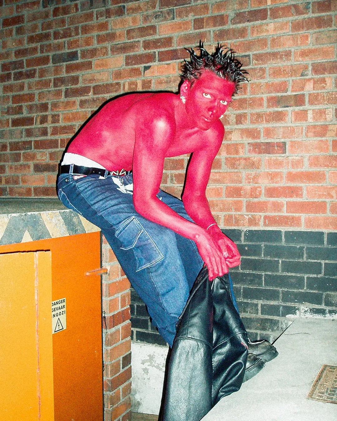 Person in pink body paint and blue jeans sitting against a brick wall.