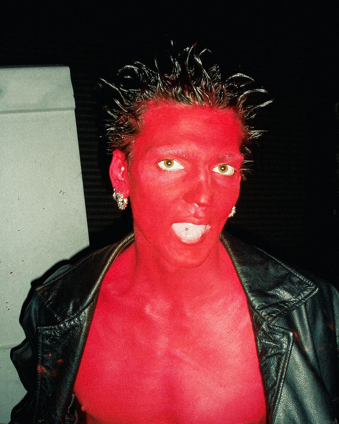 Person with red face paint wearing a black leather jacket against a dark background