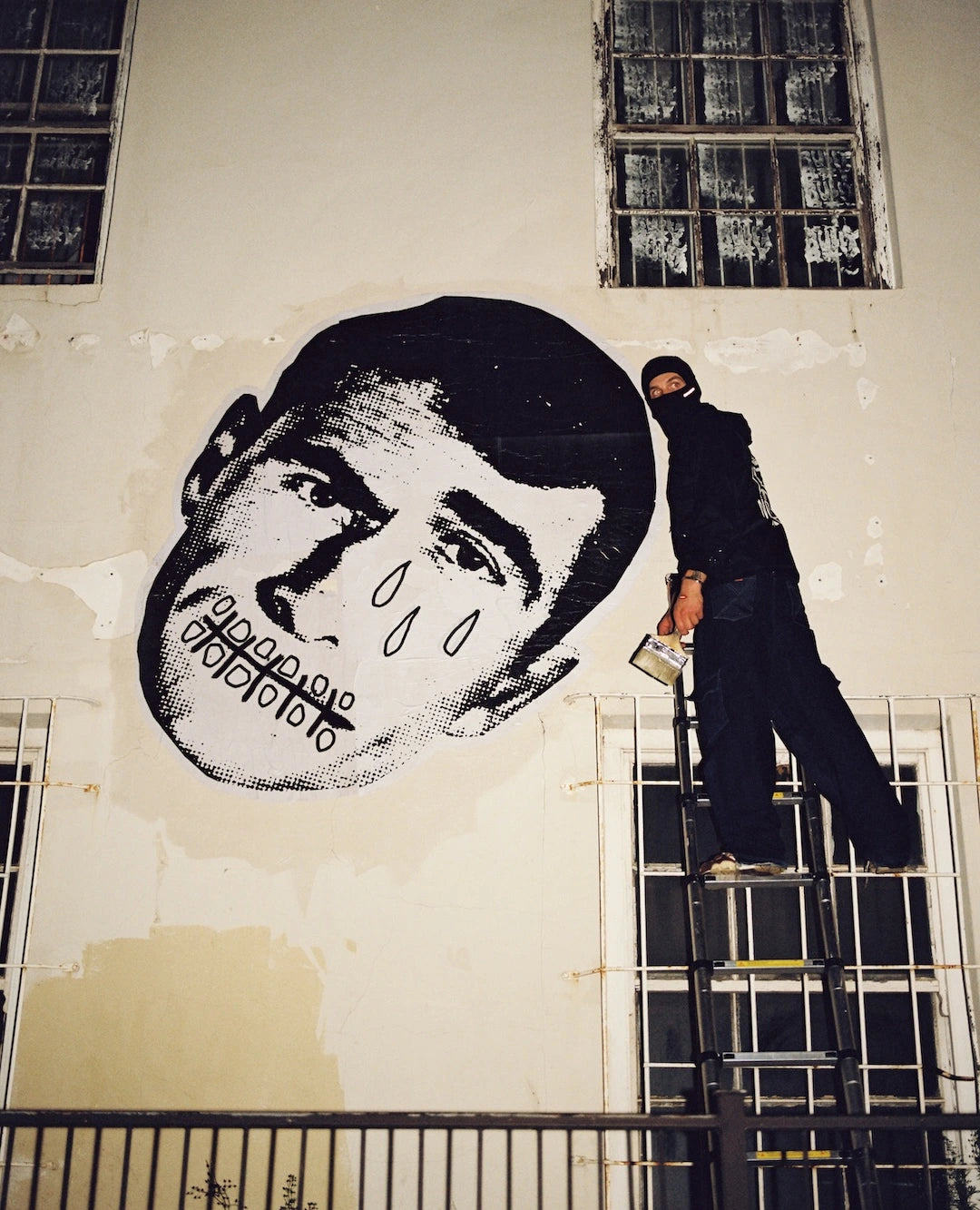 Person on a ladder painting a large mural of a face on a wall.