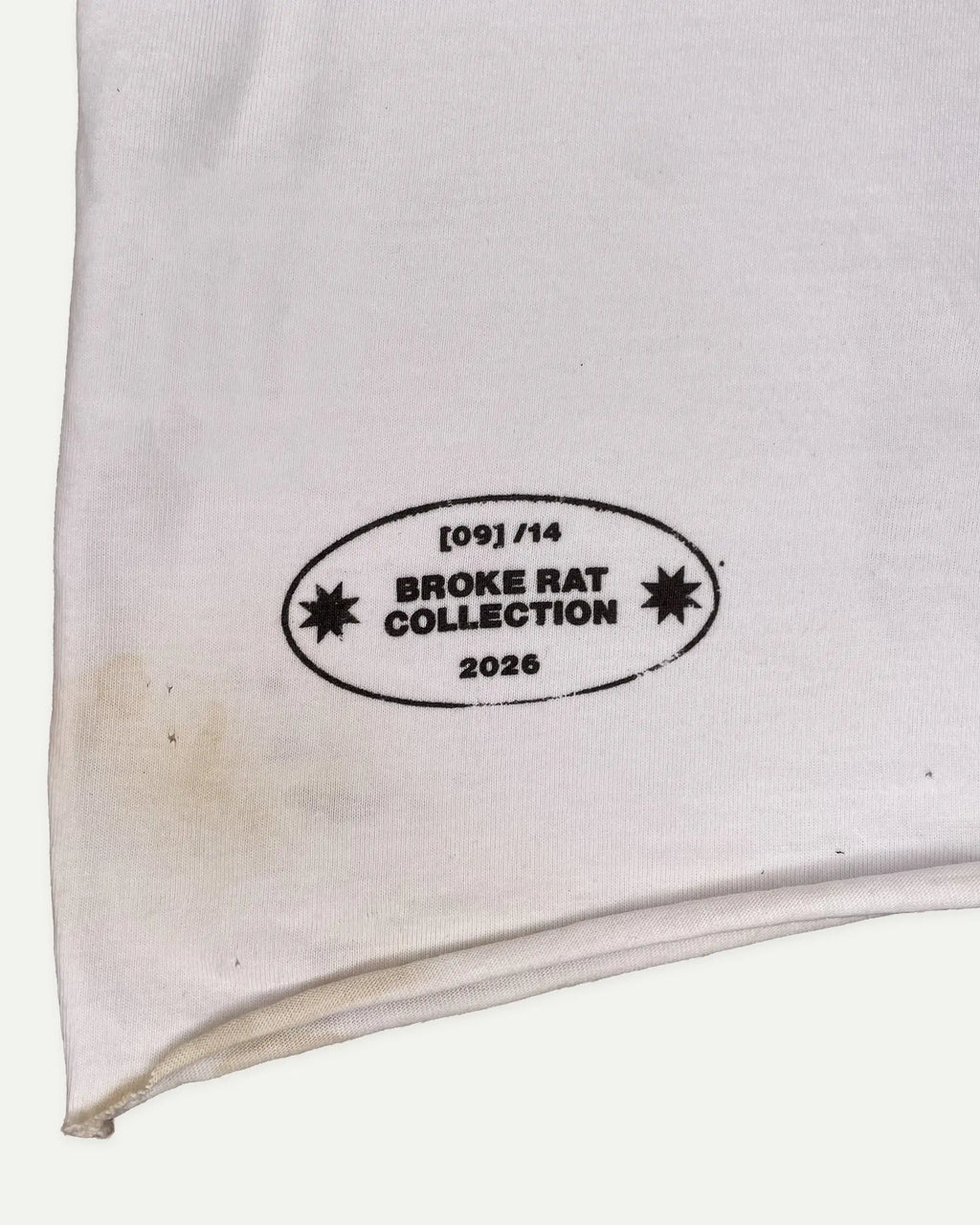 A close-up of a white fabric item, likely a sweatshirt or jacket sleeve, featuring a black printed oval label that reads '[09]/14 BROKE RAT COLLECTION 2026' with starburst icons.