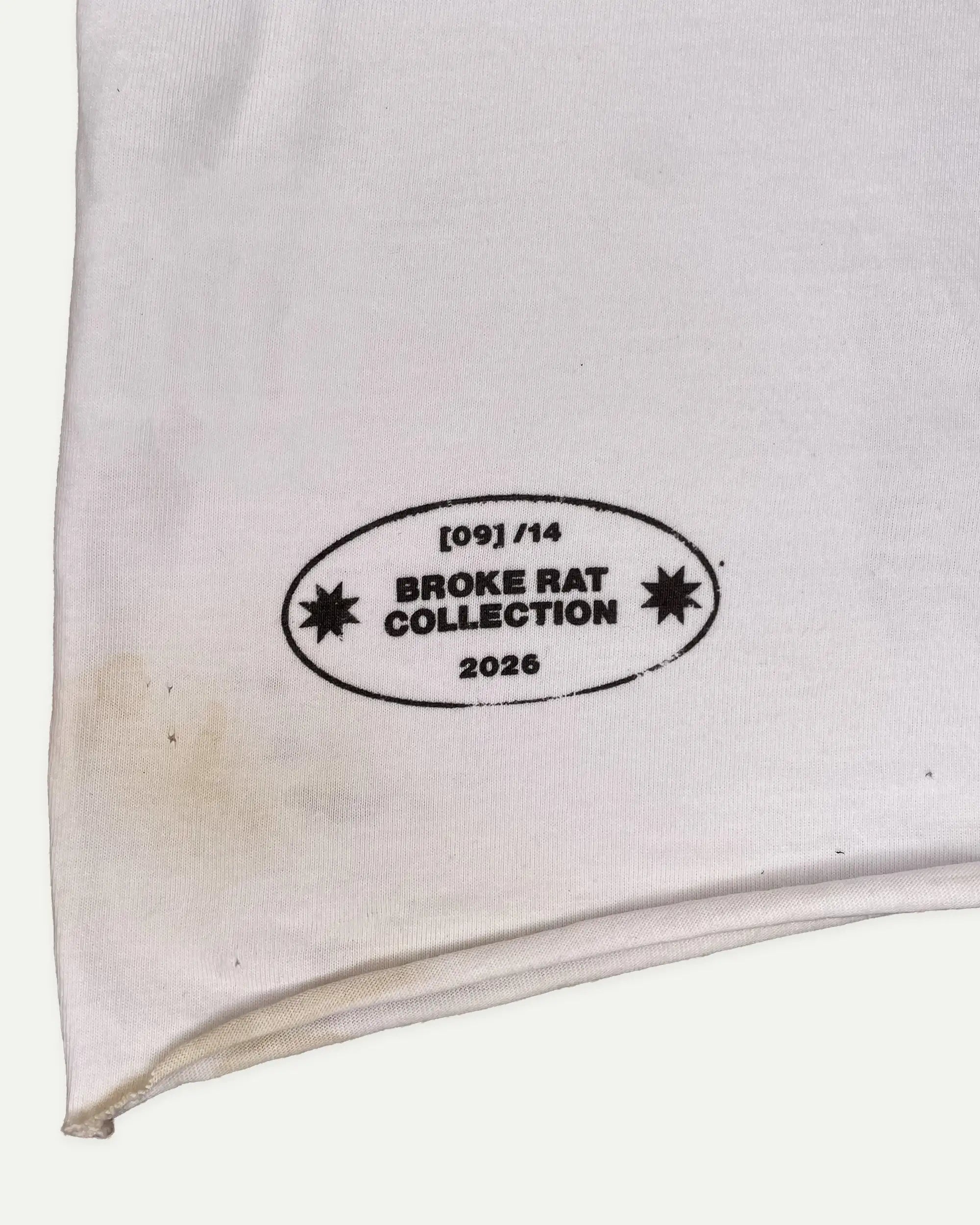 A close-up of a white fabric item, likely a sweatshirt or jacket sleeve, featuring a black printed oval label that reads '[09]/14 BROKE RAT COLLECTION 2026' with starburst icons.