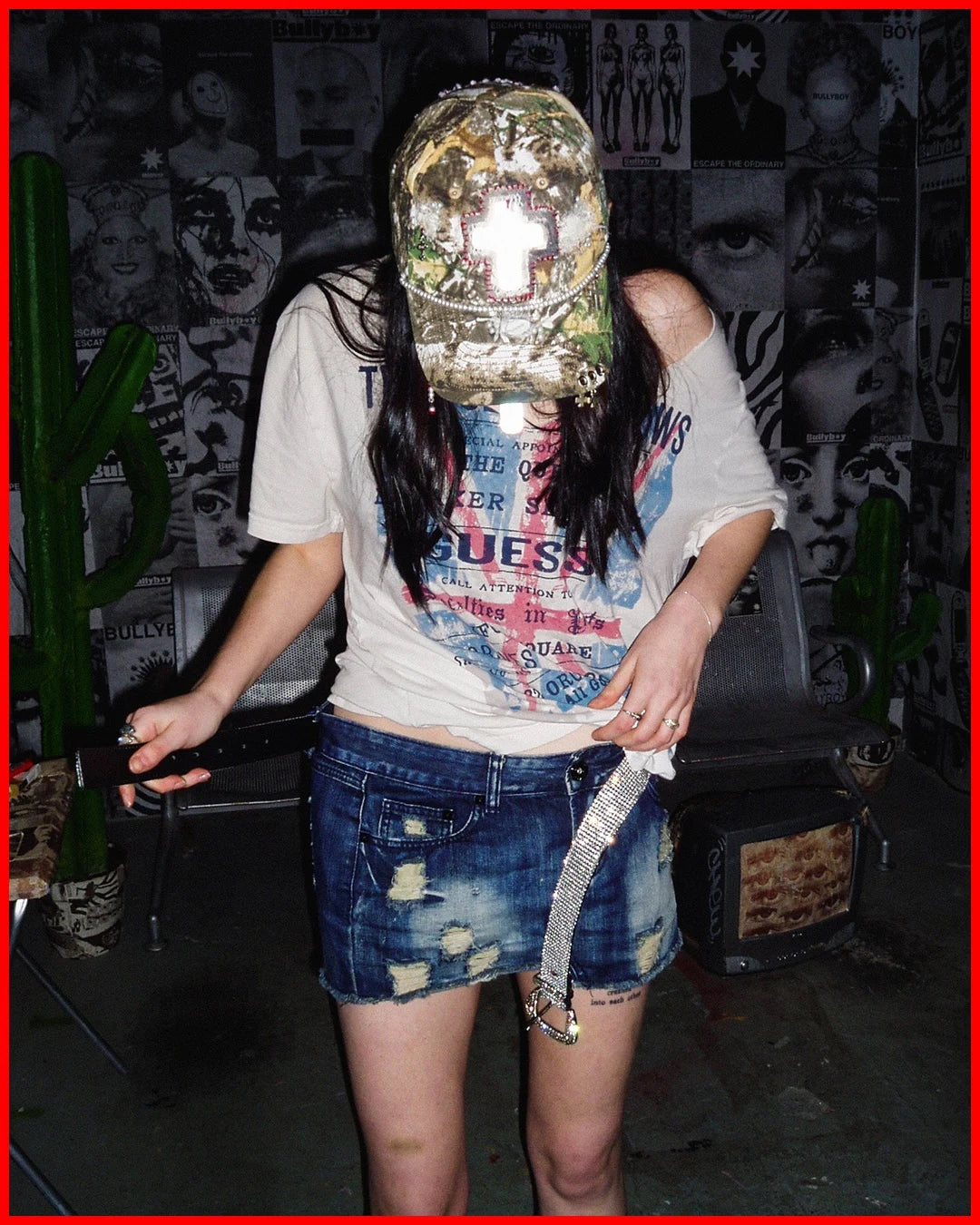 Person wearing a sequin mask and Guess t-shirt in a room with a cactus plant and graffiti wall.