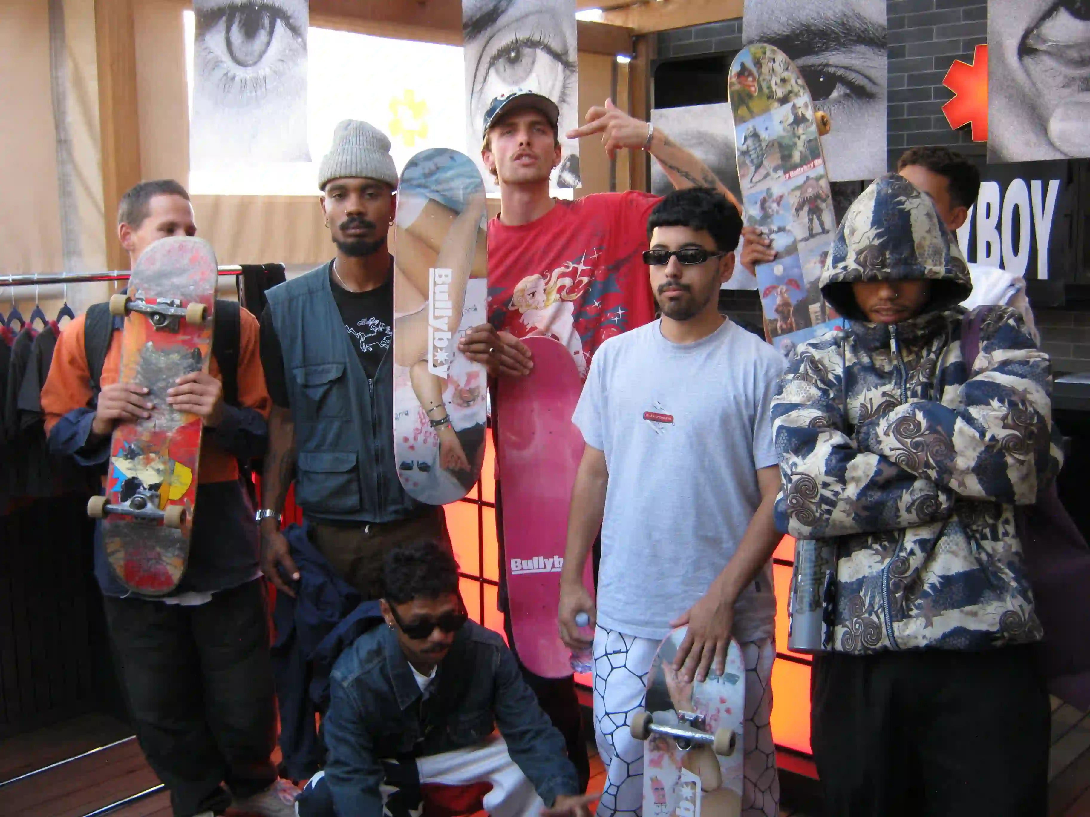 Group of six men with skateboards posing confidently in casual attire. Background features artistic eye posters and an urban setting. Vibrant and energetic vibe.