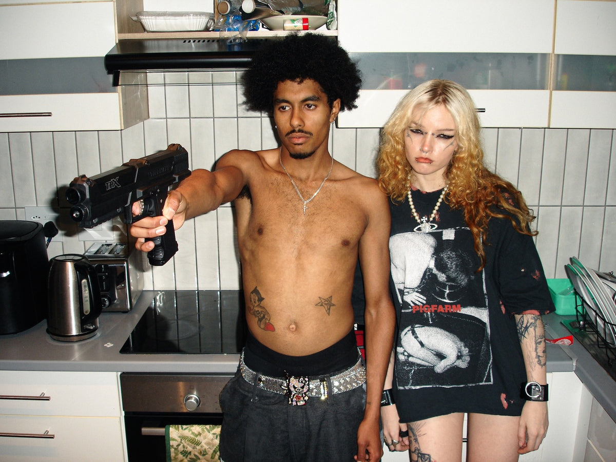 A shirtless man with an afro holding a black handgun points it toward the camera, standing next to a blonde woman in a kitchen setting.
