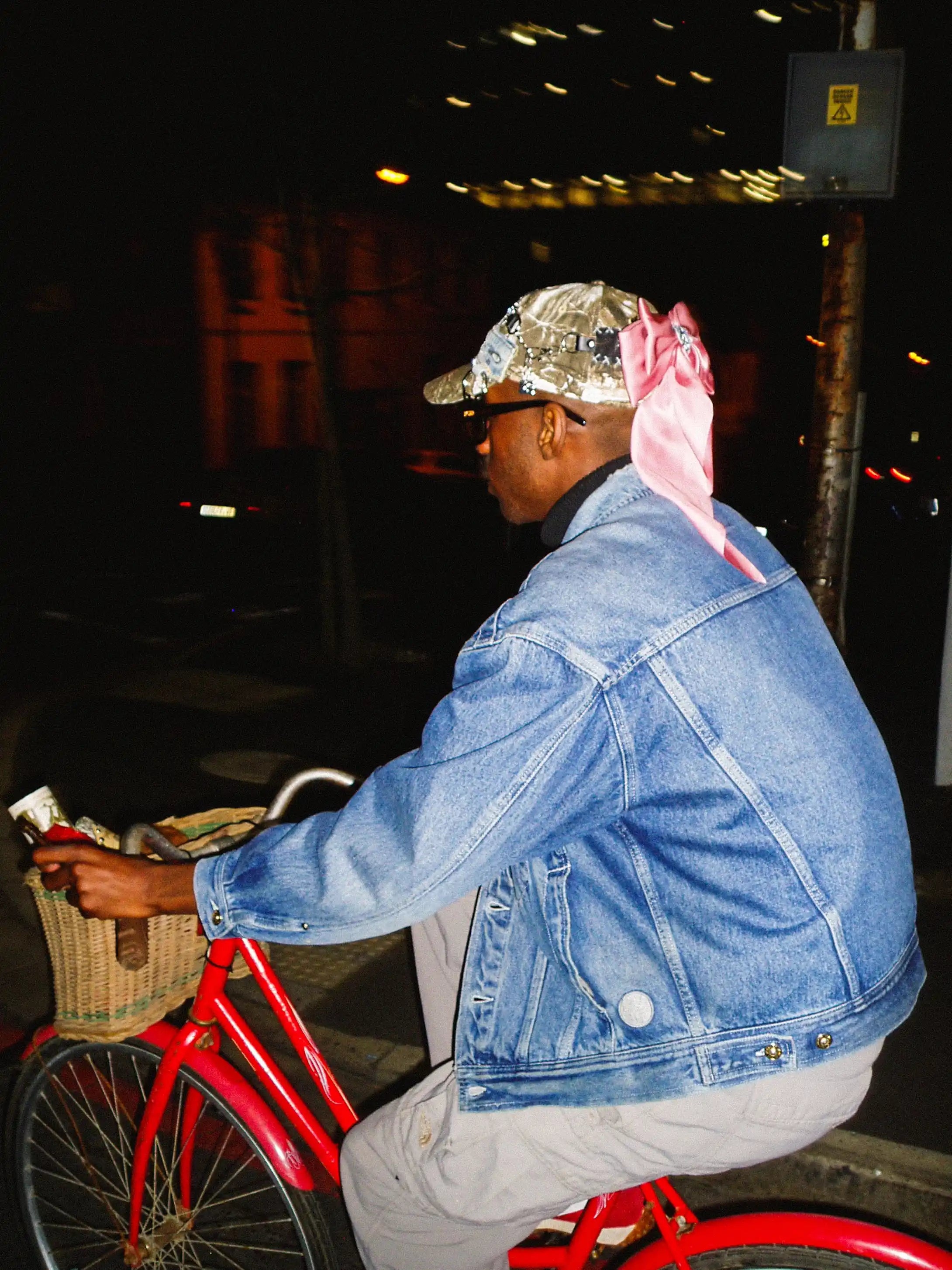 Person wearing a denim jacket and gold cap with a red bicycle at night.