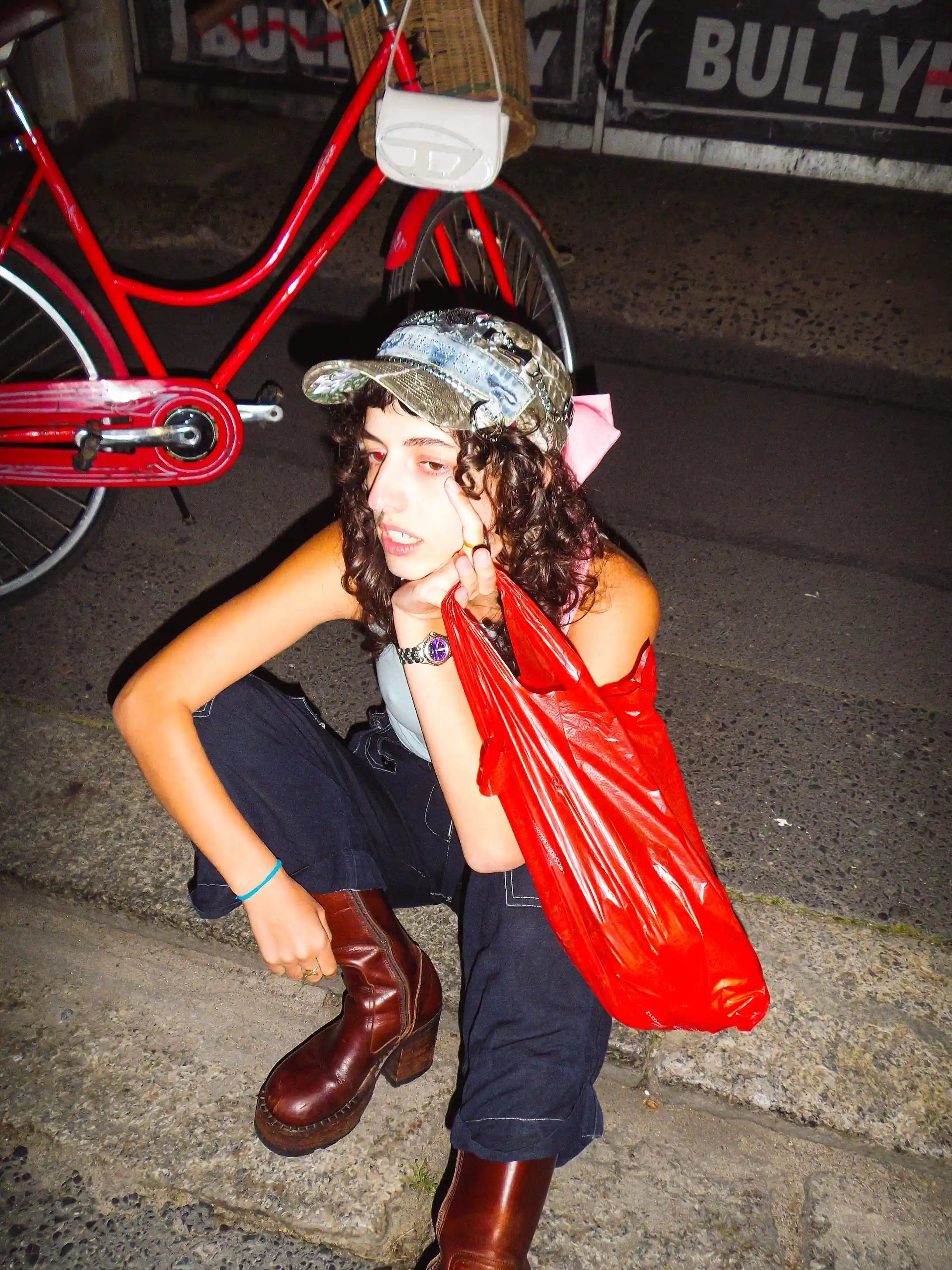 Person sitting on a curb with a red plastic bag, wearing a cap and boots, with a red bicycle and 'BULLY' sign in the background.