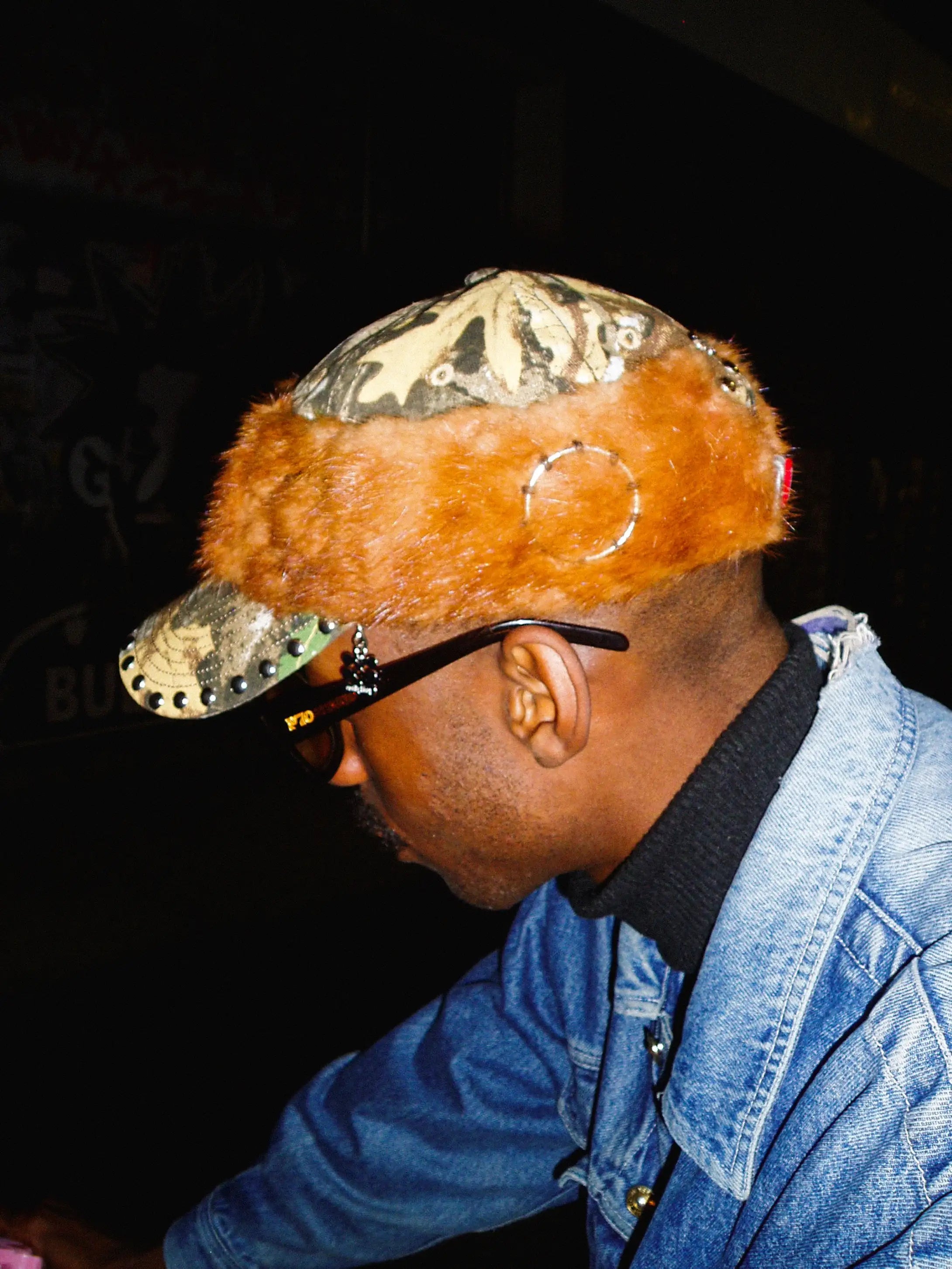 Person wearing a unique hat with camouflage and fur design, sunglasses, and a denim jacket.