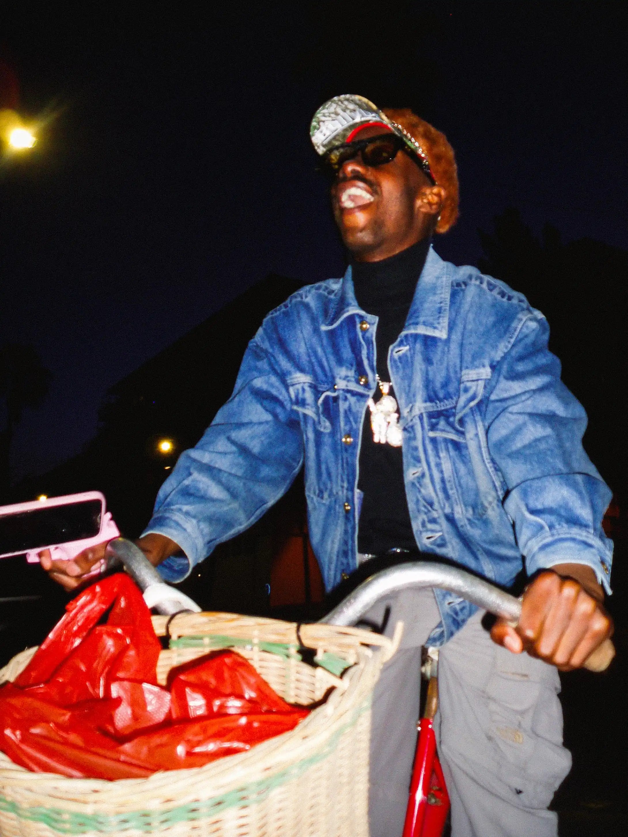 Person pushing a bicycle with a basket containing red bags at night.