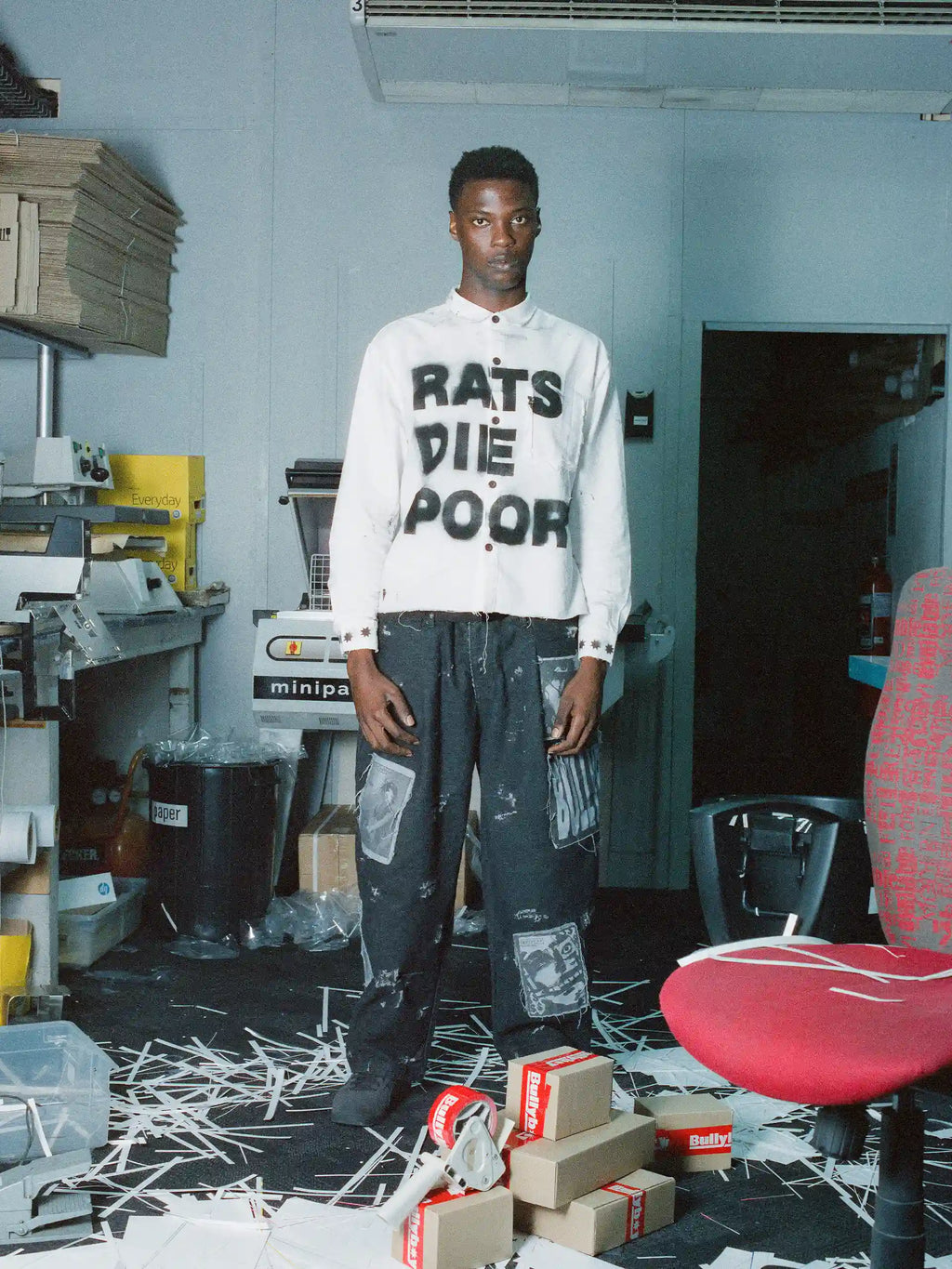 A man stands in a cluttered indoor workspace wearing a white long-sleeve shirt with bold black text reading 'RATS DIE POOR' and dark distressed pants with graphic patches. Shredded paper covers the floor, with cardboard boxes and a red office chair visible around him.