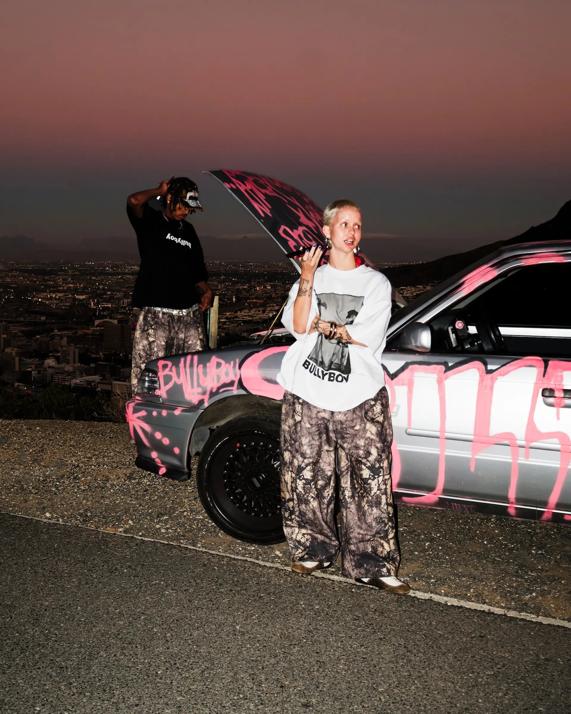 Two people standing next to a car with graffiti at dusk.