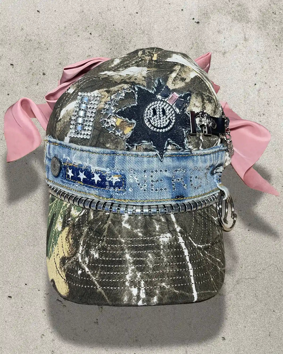 Decorative cap with pink ribbons, embellishments, and a camouflage pattern on a concrete surface.
