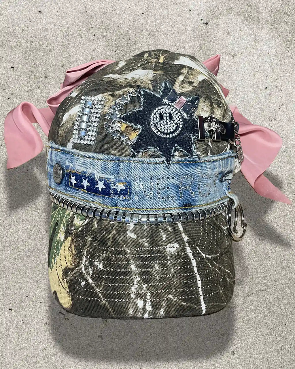 Decorative cap with pink ribbons, embellishments, and a camouflage pattern on a concrete surface.