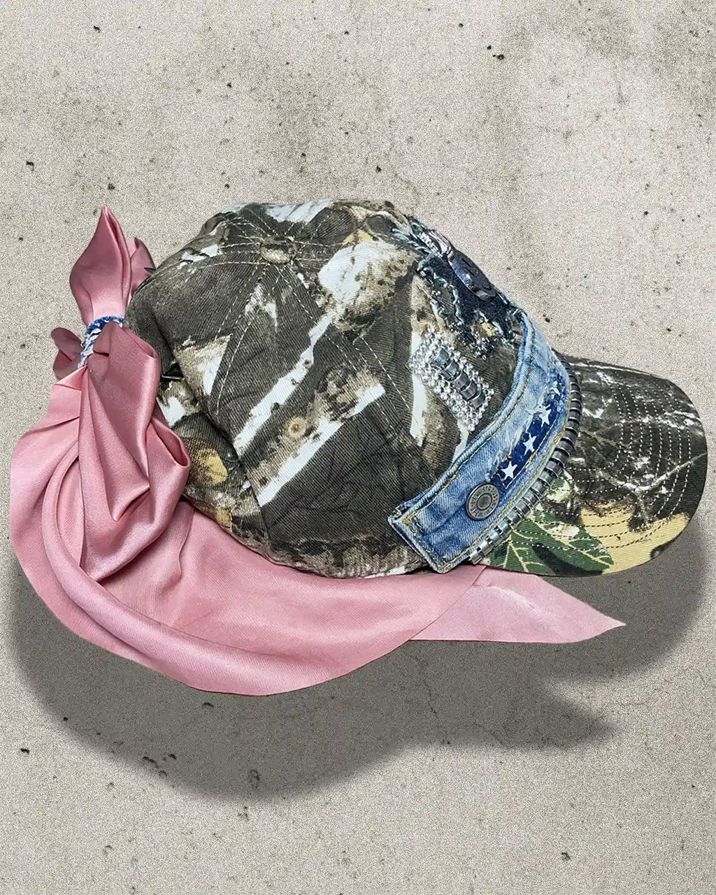 Camouflage cap with denim brim and pink scarf on a concrete surface