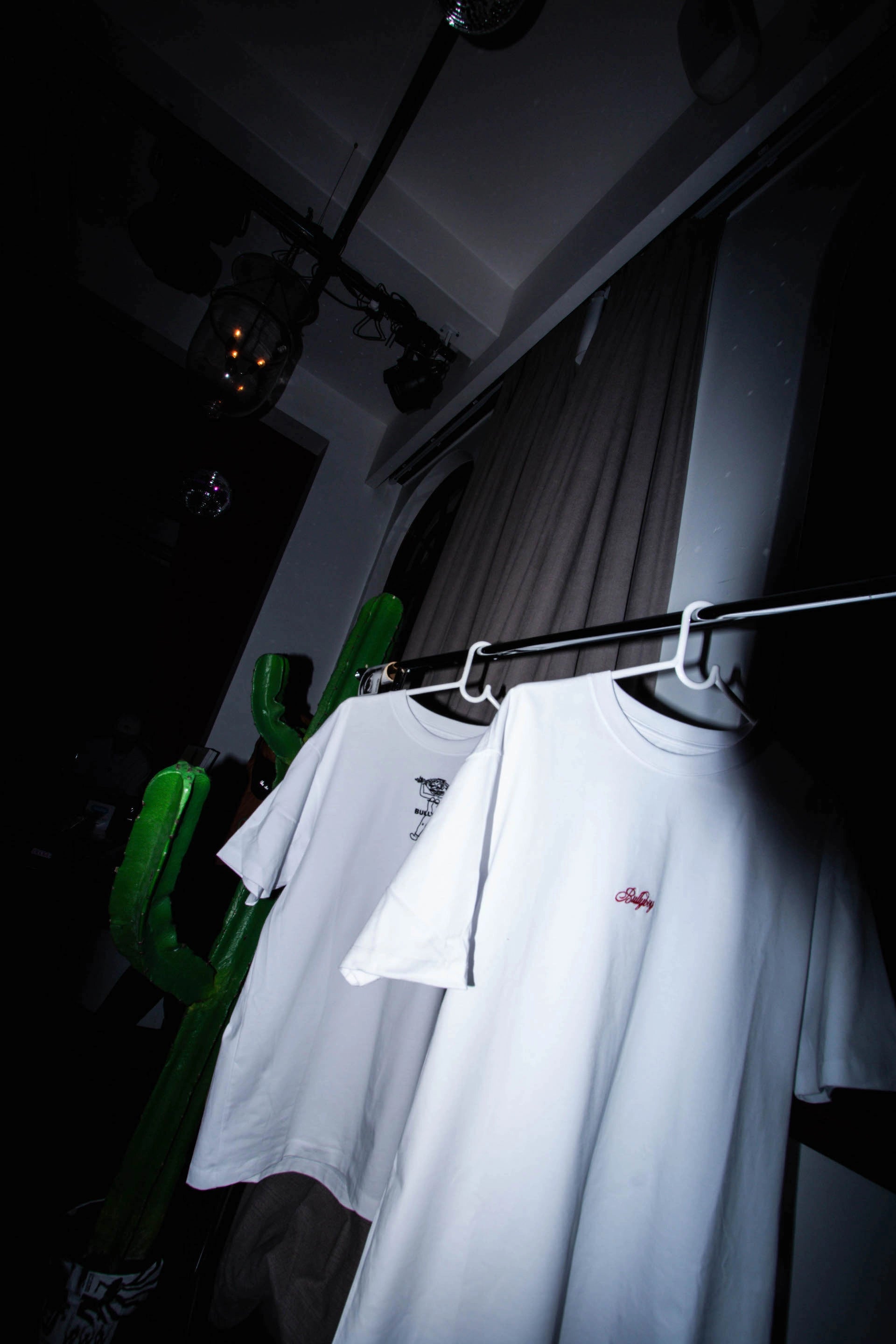 A pristine white t-shirt elegantly displayed on a rack in a dimly lit room at the Buzzcut market, Gorgeous George hotel