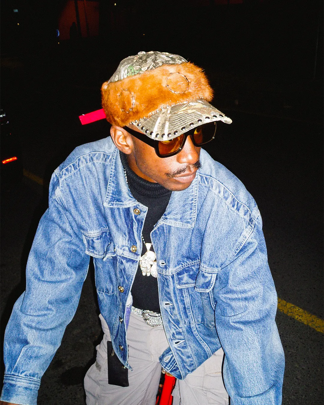 Person wearing a denim jacket, sunglasses, and a cap with a dark background