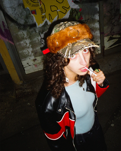 Person wearing a camouflage hat with a fur-like top, black leather jacket with red accents, and light blue shirt in a dark setting.