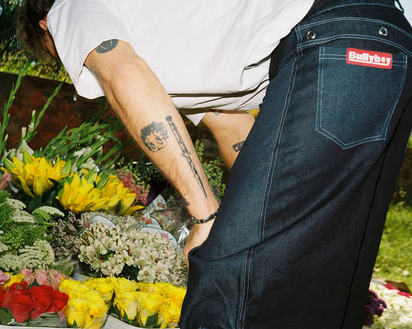 A man with a tattoo on his arm bends over a vibrant flower arrangement, showcasing streetwear style from Bullyboy Skateshop